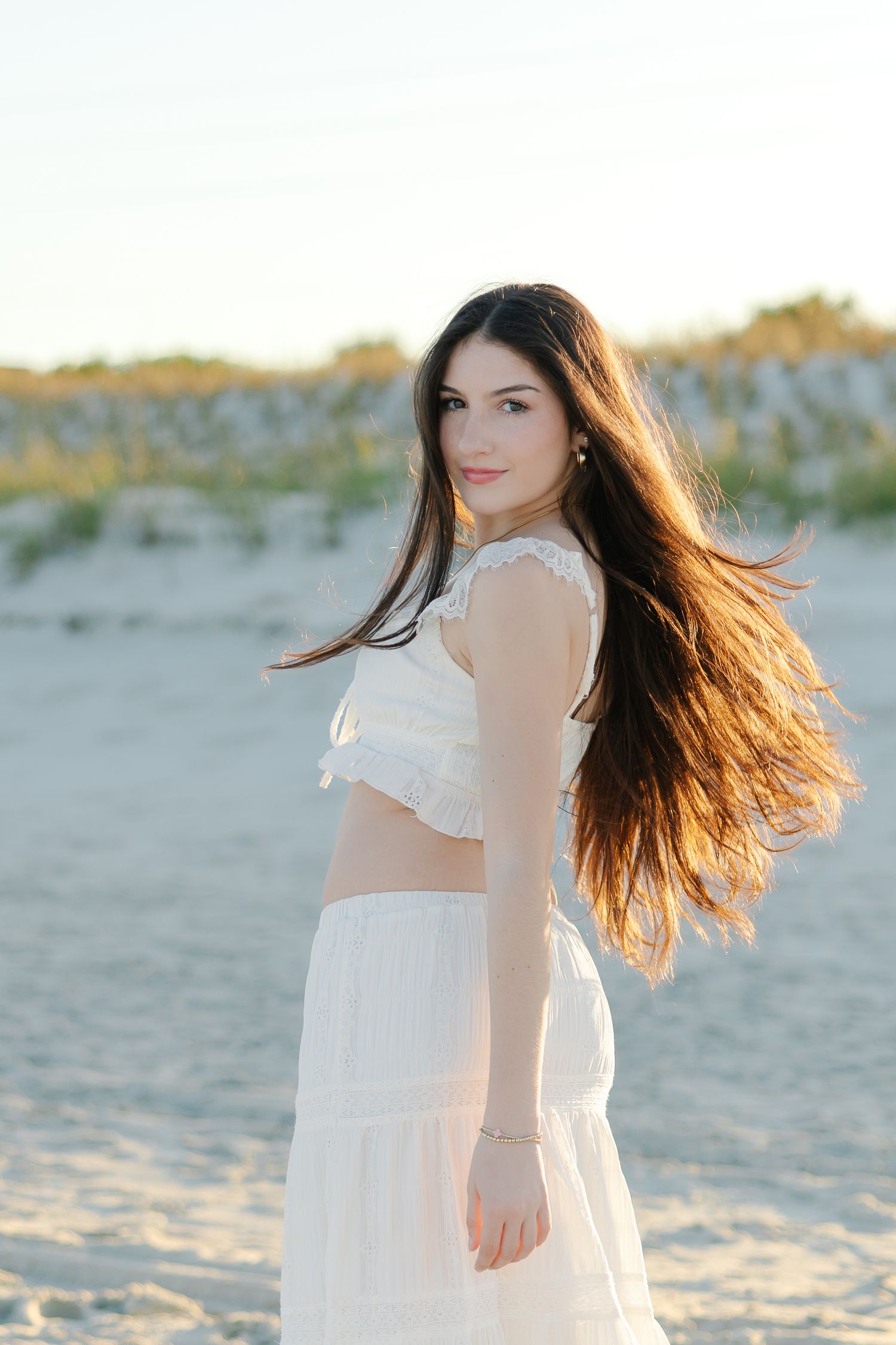 Young woman in white crop top and skirt with flowing hair posing on sandy beach.