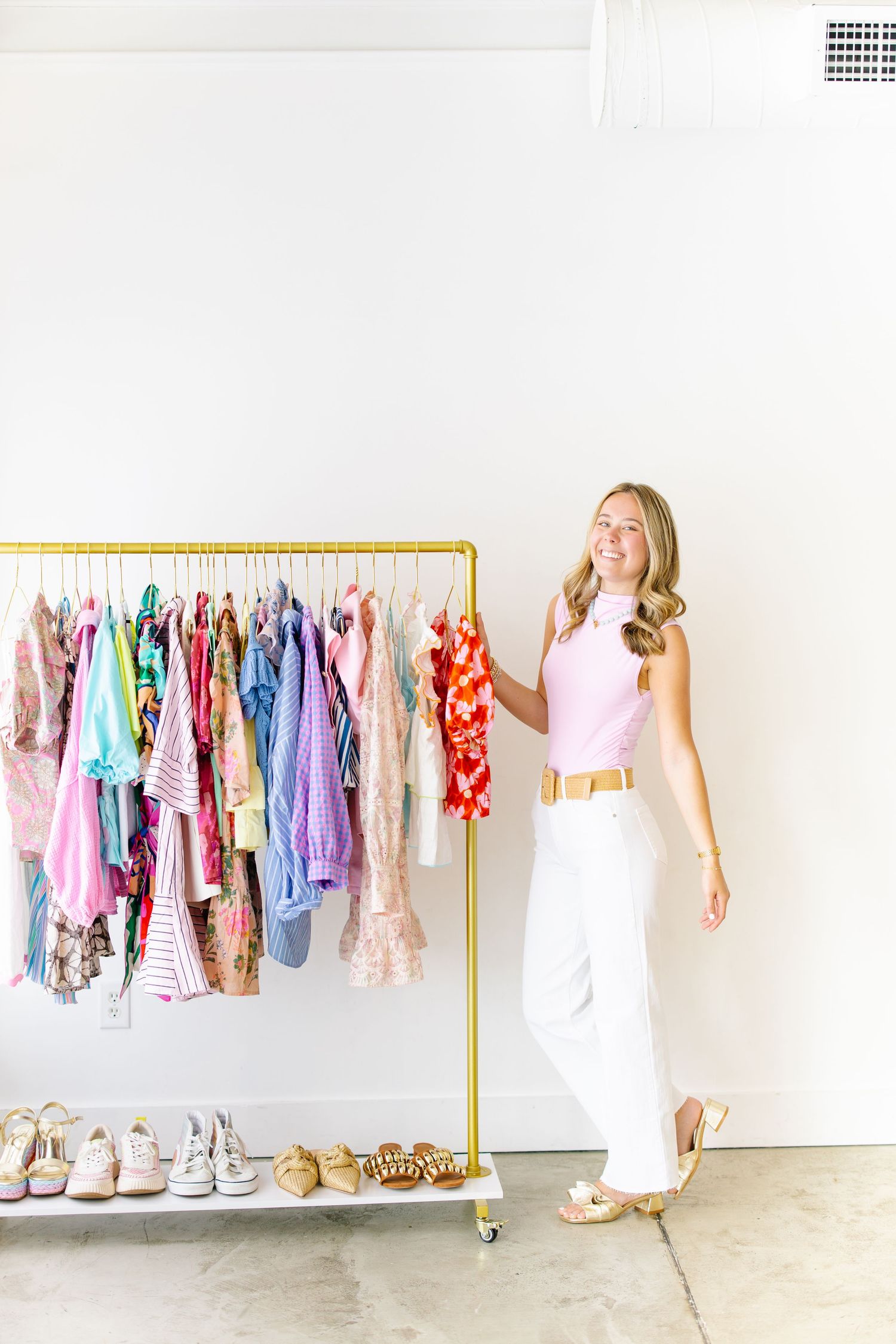 Blonde woman in pink sleeveless top and white pants browsing colorful clothes on a gold rolling rack.