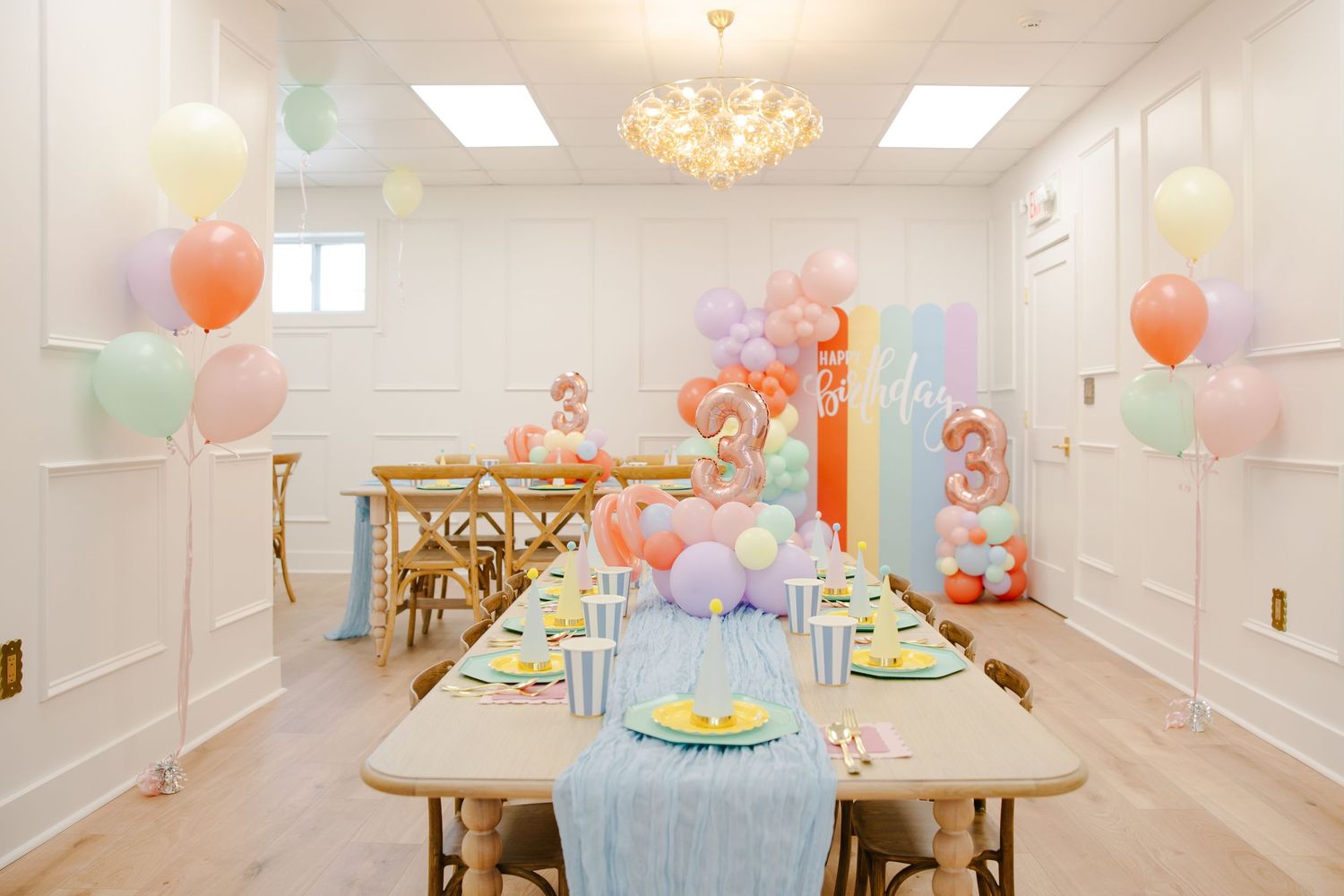 Elegant children's birthday party room with pastel balloon decor, blue table runner, and chandelier lighting.