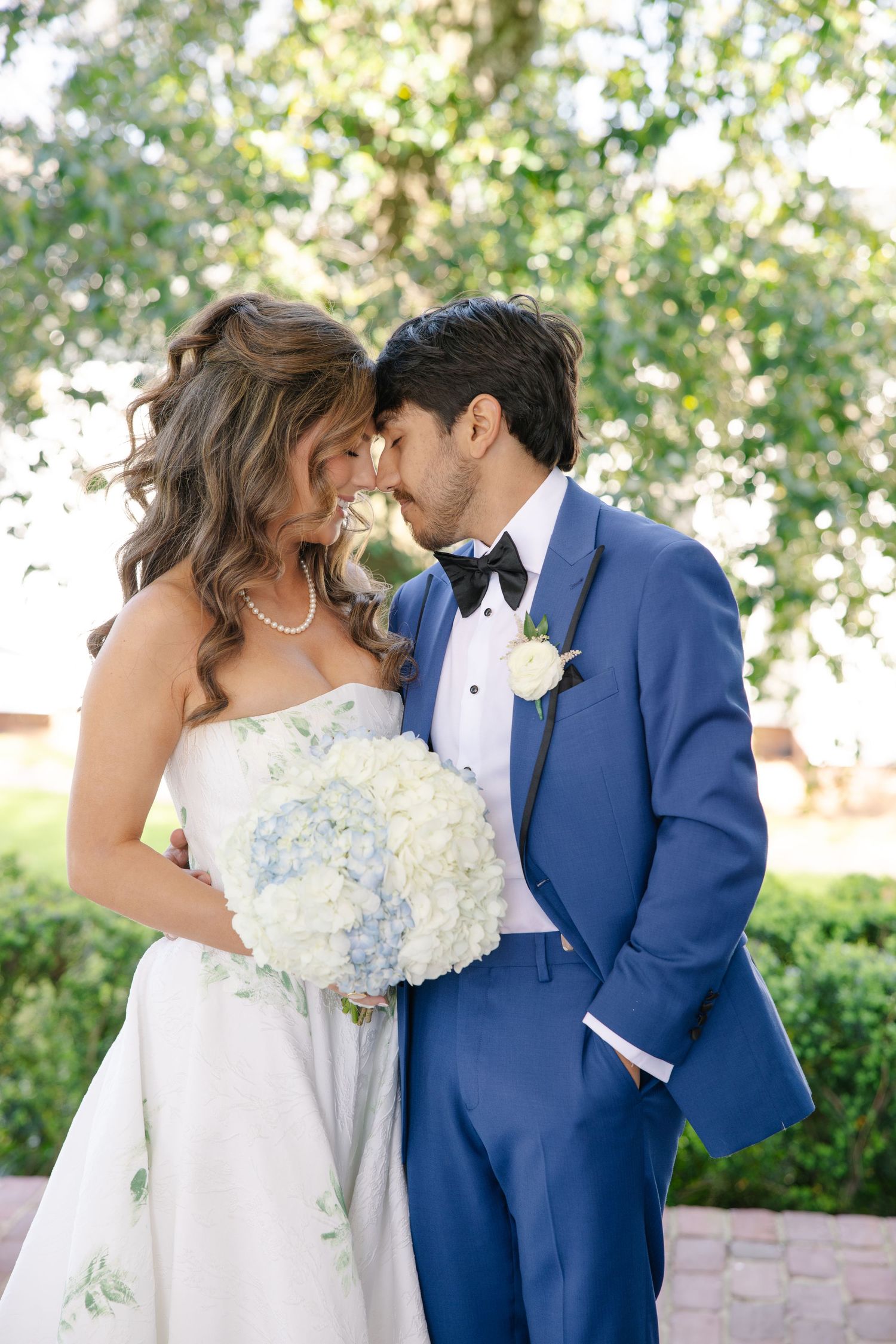 Bride and groom touch foreheads tenderly as she holds a blue hydrangea bouquet, groom in a blue tuxedo suit.