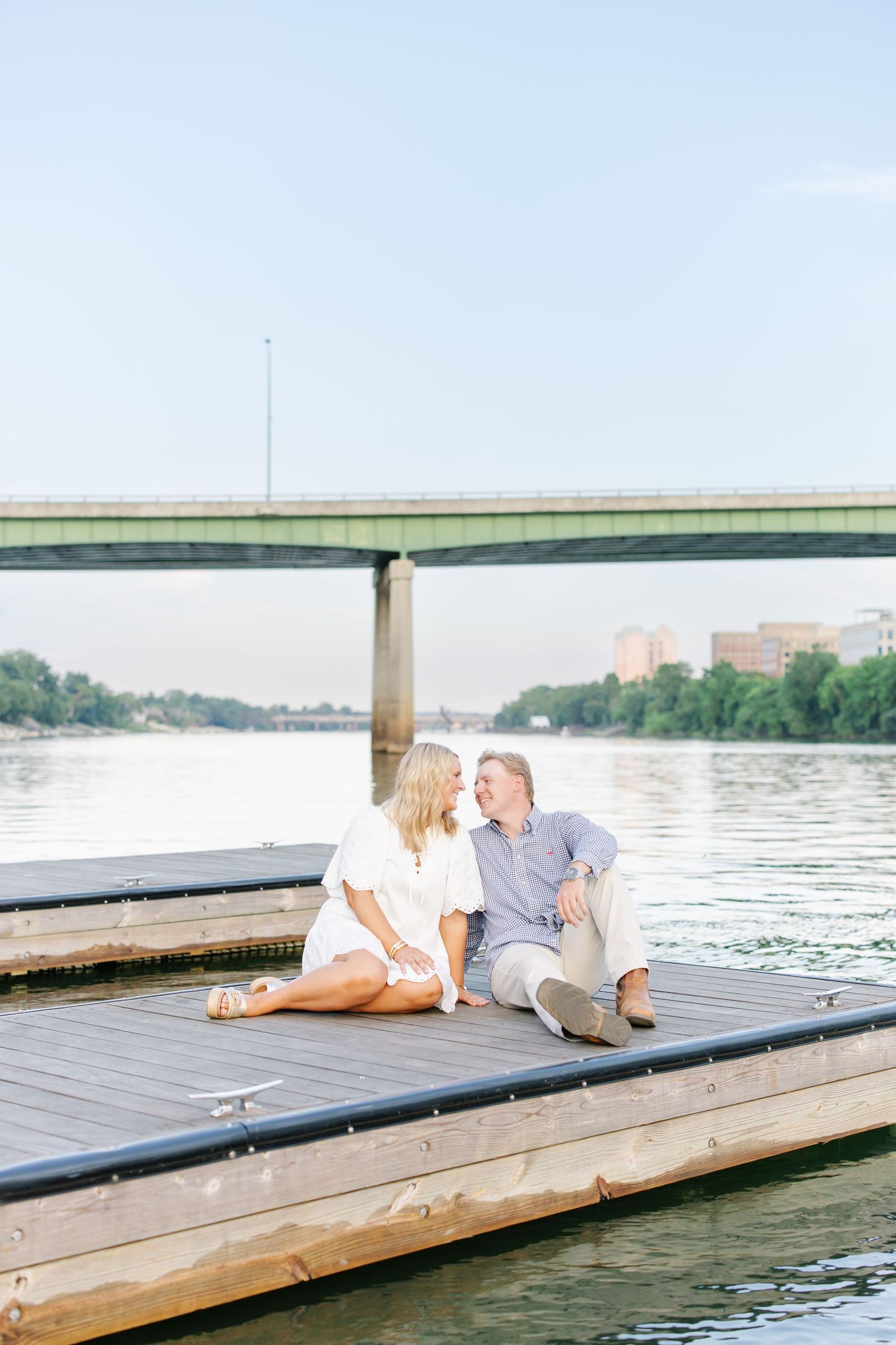 Couple sitting together on wooden dock by river with bridge in background.