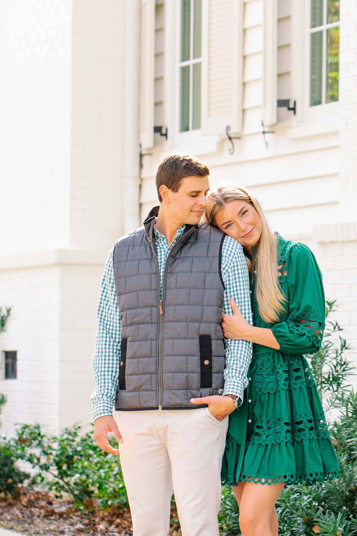 Couple posing outdoors, woman in green dress leaning on man in gray vest and plaid shirt near white building.