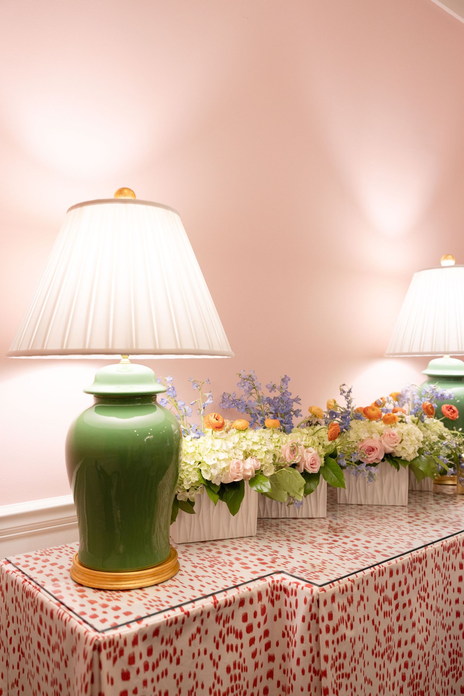 Green ceramic ginger jar lamps flanking fresh floral arrangements on a pink spotted tablecloth against a pink wall.