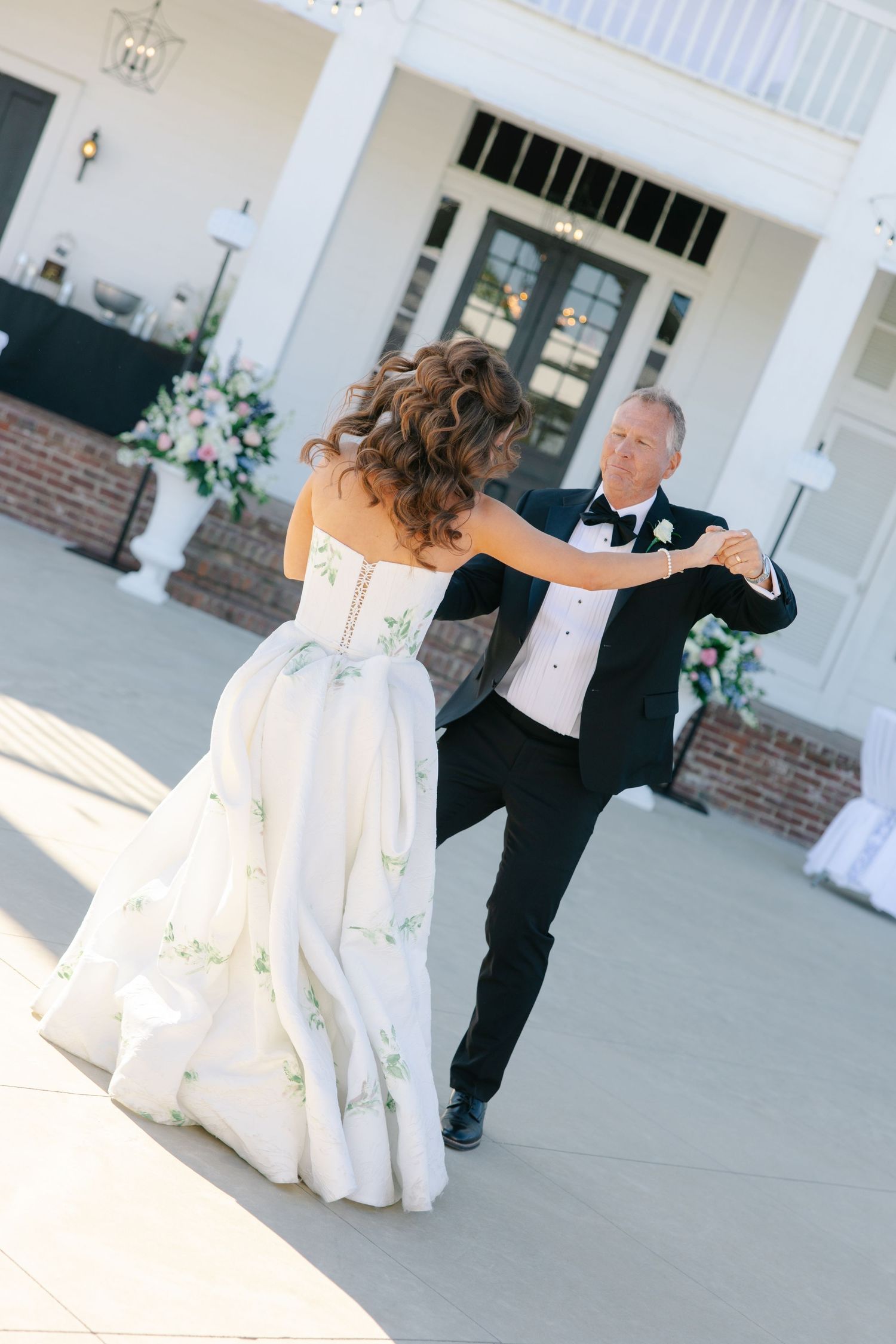 Bride in floral white gown dancing with older man in tuxedo at outdoor wedding reception.