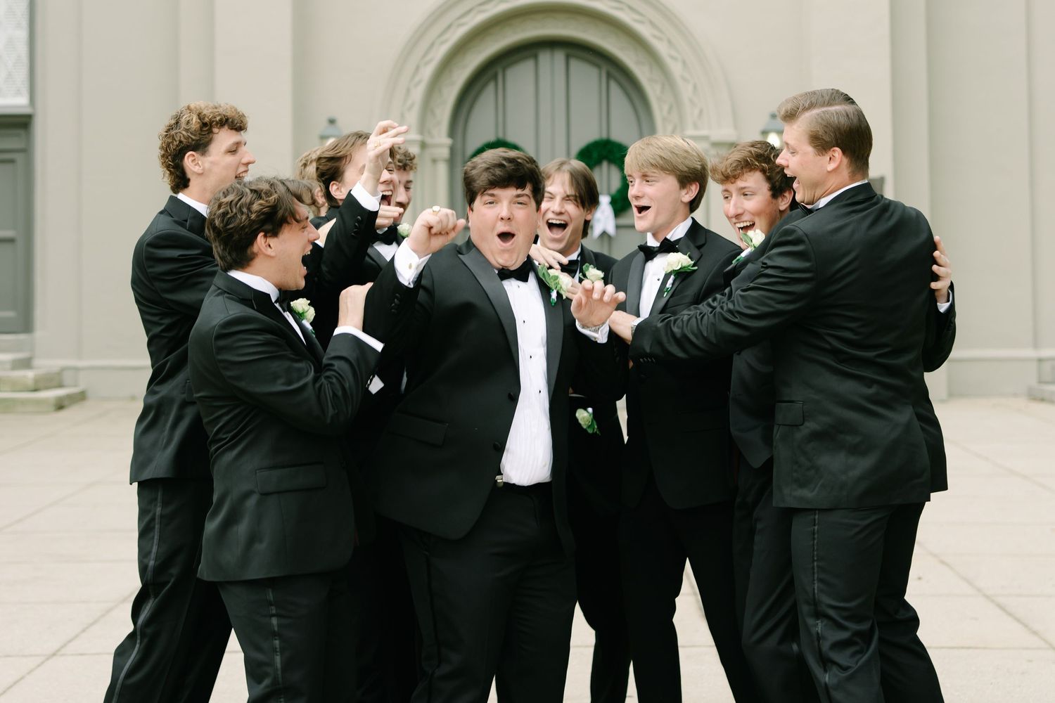 Group of young men in black tuxedos with boutonnieres celebrating together outside a church.