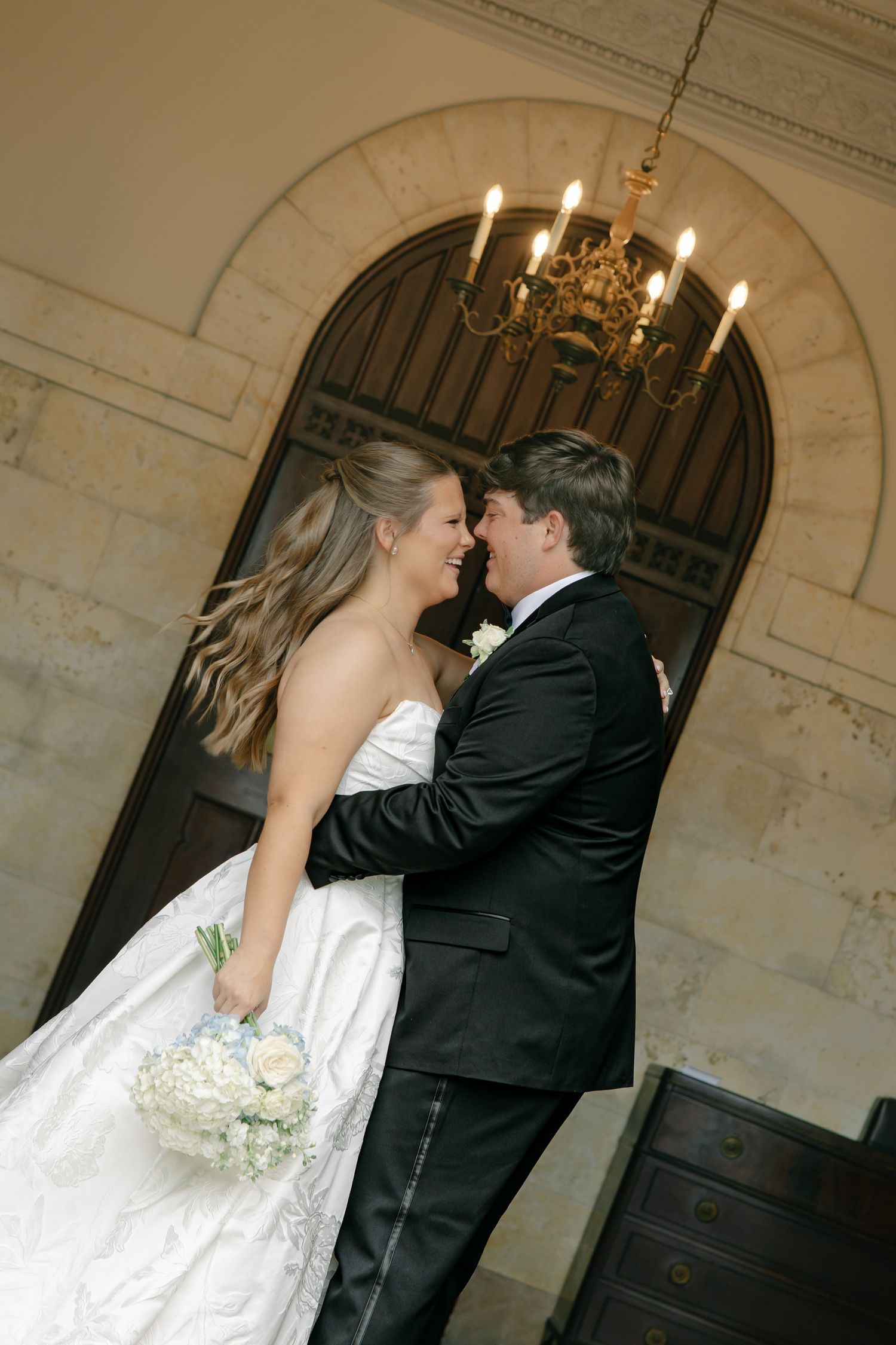 Bride and groom share a romantic moment in elegant venue, bride holding white bouquet in strapless gown, groom in black tuxedo.