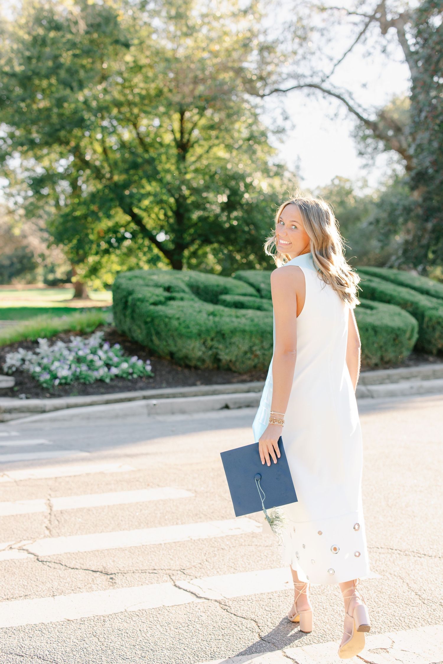 Smiling blonde woman in white dress holding graduation cap while crossing a sunny street.