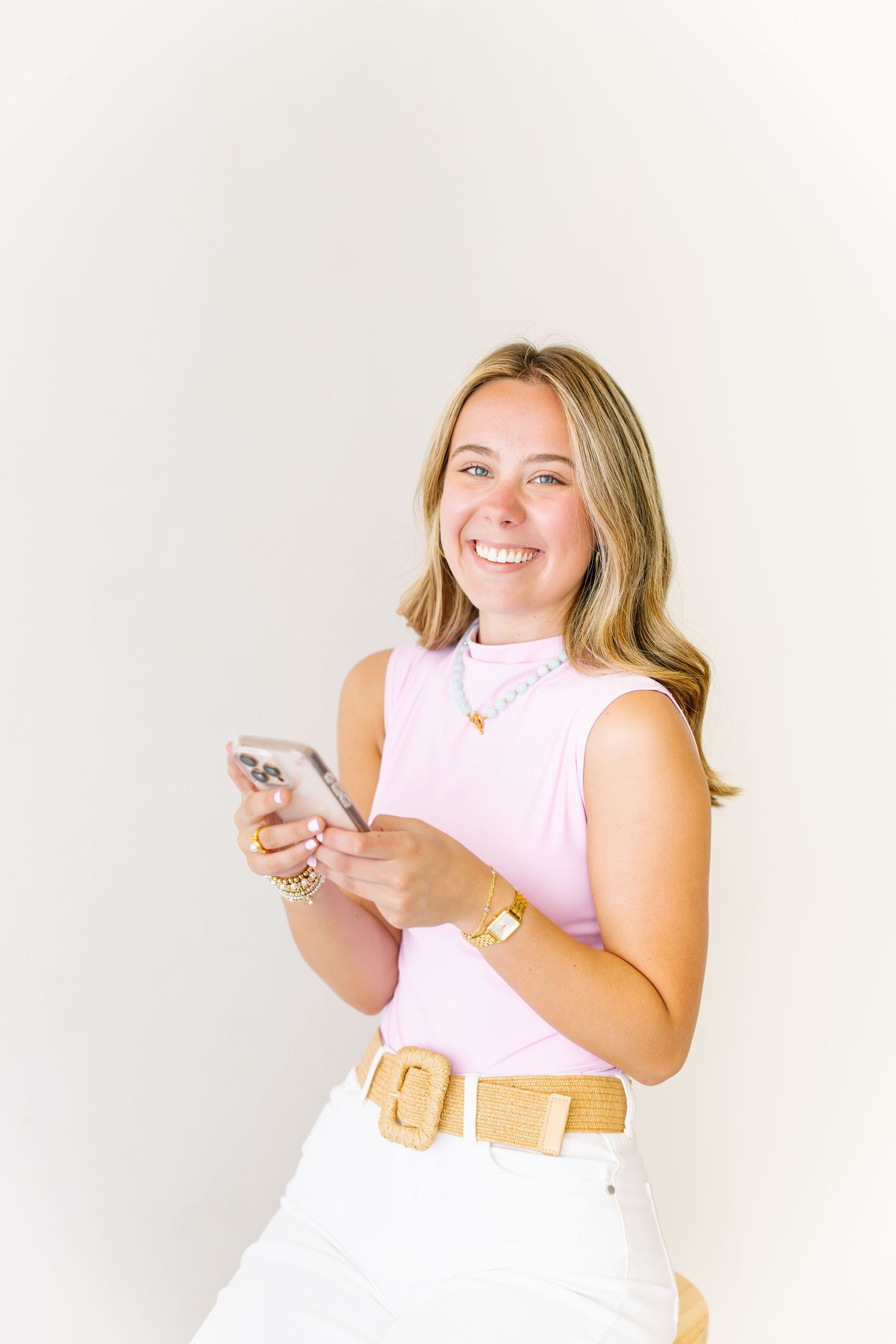 Smiling woman in pink top and white pants holding a smartphone while wearing gold accessories.