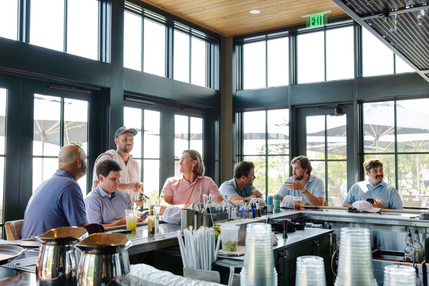 Group of businessmen gathered around a bar counter in a modern clubhouse with large windows.