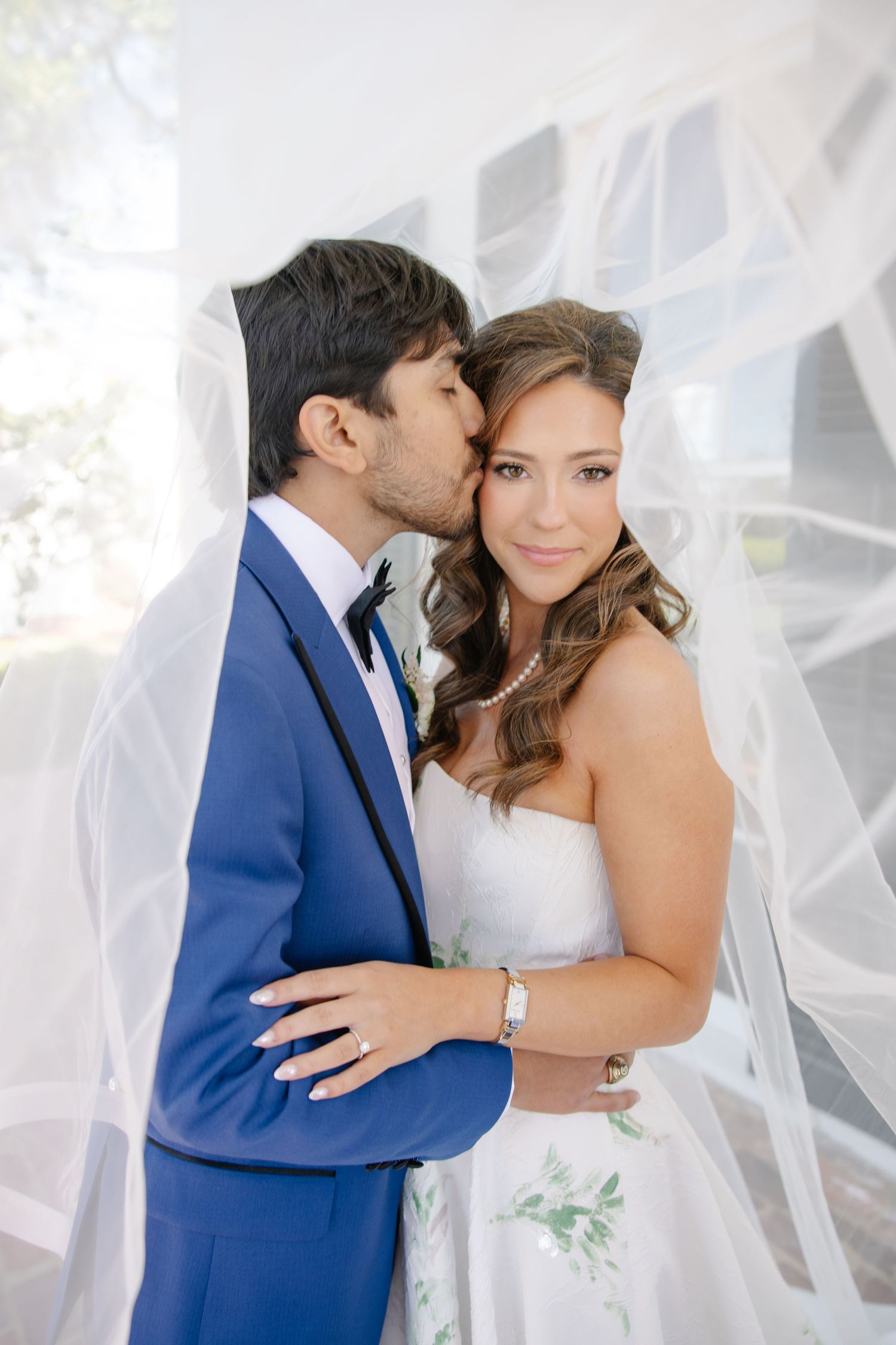 Groom in blue suit kissing bride on cheek under a white veil at outdoor wedding.