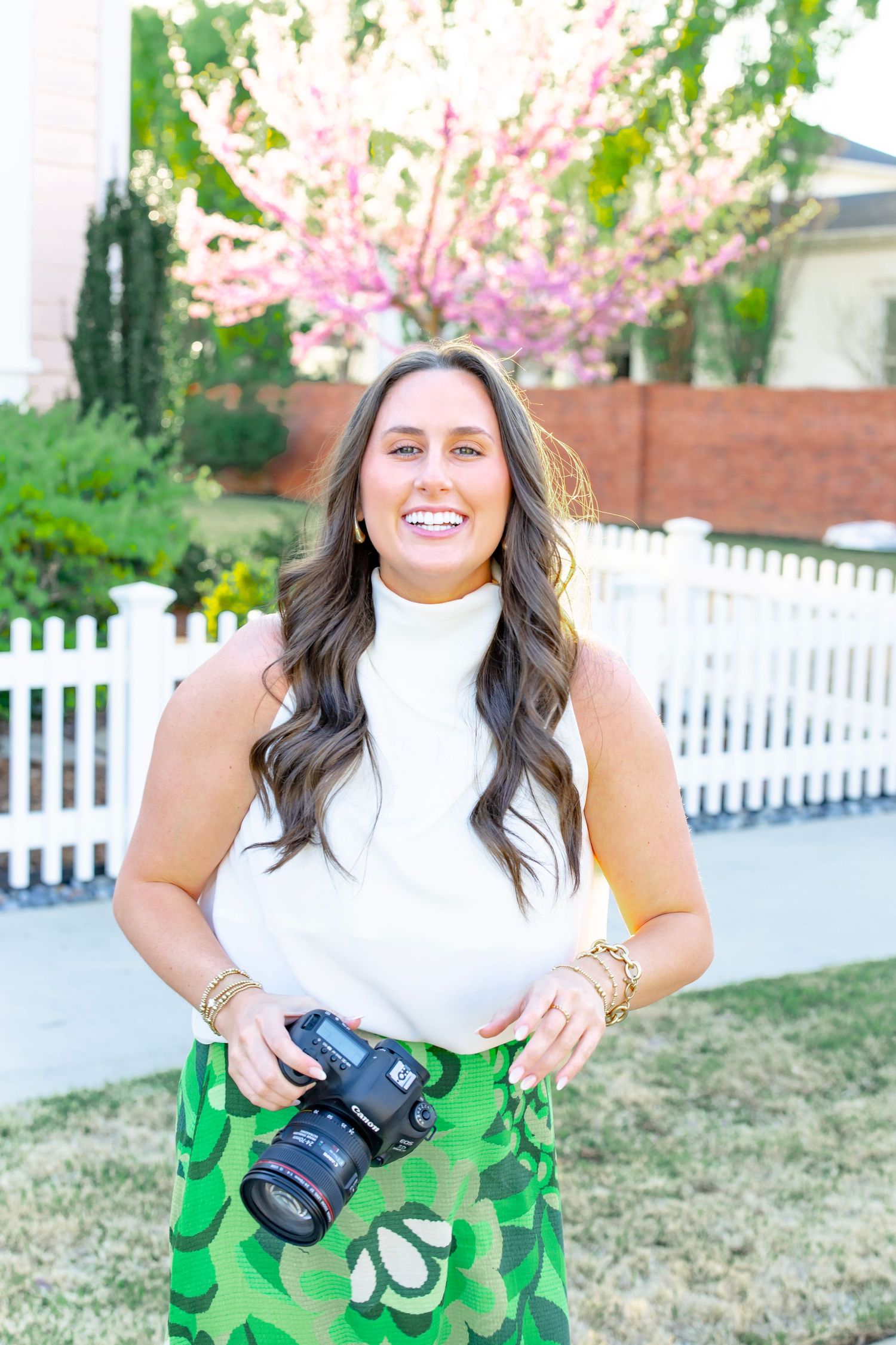 Smiling brunette photographer holding a camera outdoors near a white fence with pink flowering trees behind her.