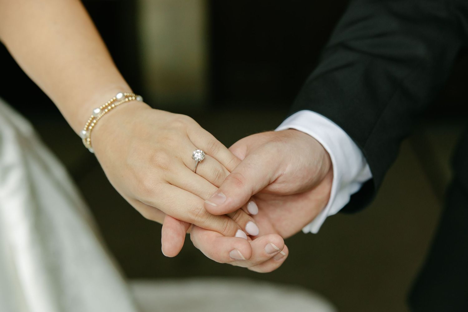 Bride and groom holding hands, showcasing diamond engagement ring and pearl bracelet on wedding day.
