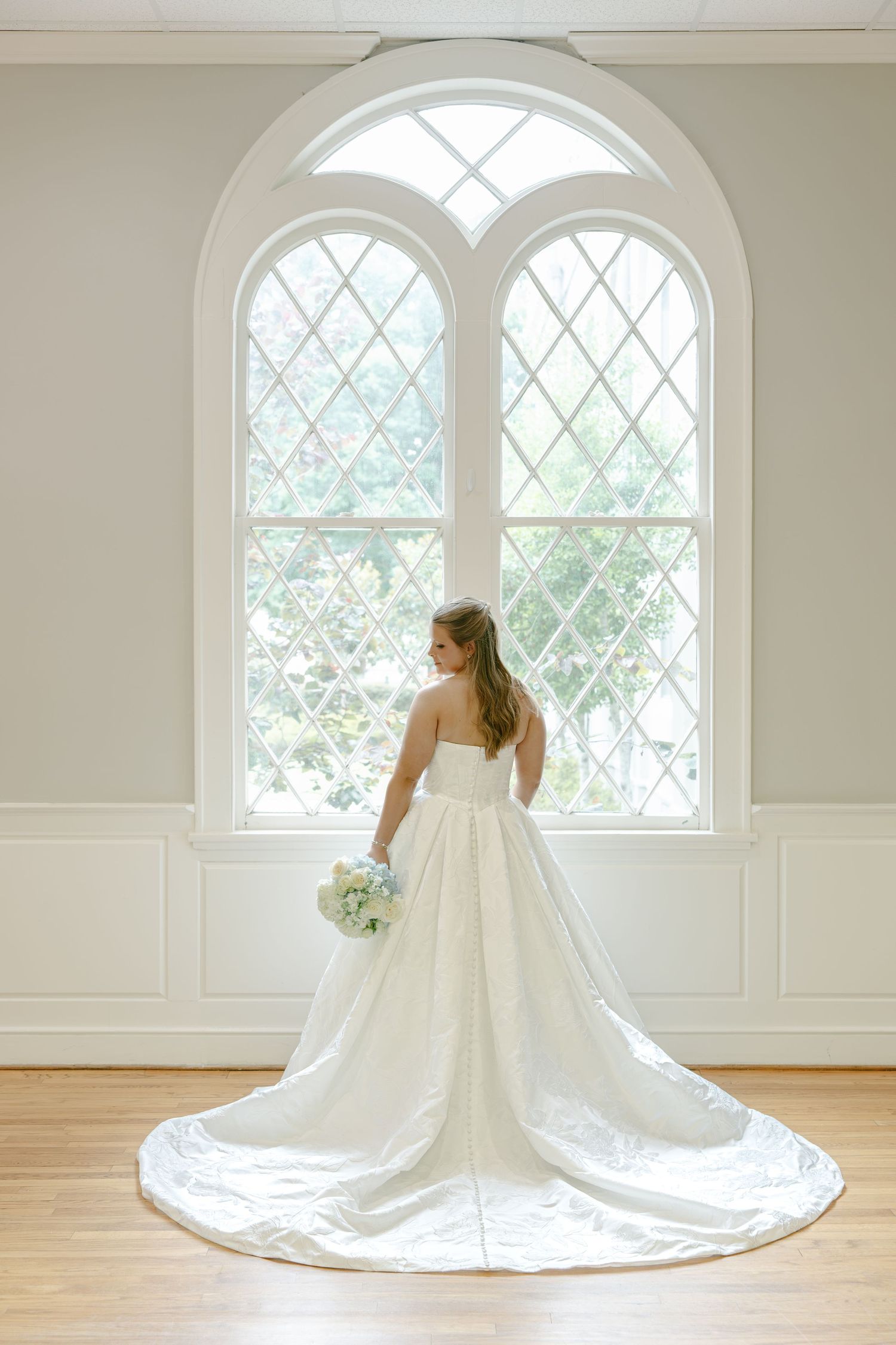 Bride in strapless white ball gown with long train stands before arched leaded glass window holding bouquet.