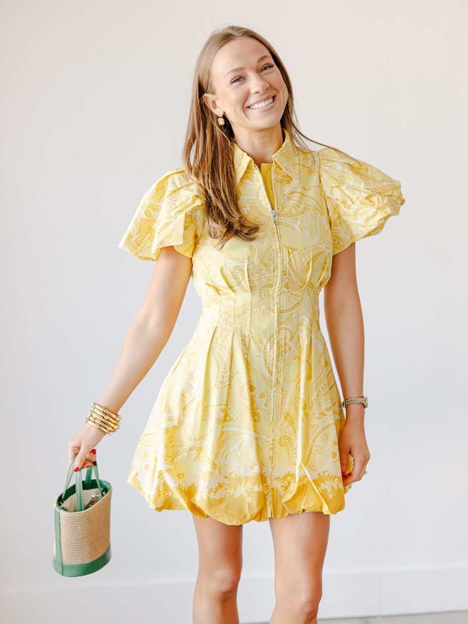 Woman wearing a yellow floral print mini dress with flutter sleeves and collar, holding a woven bucket bag.