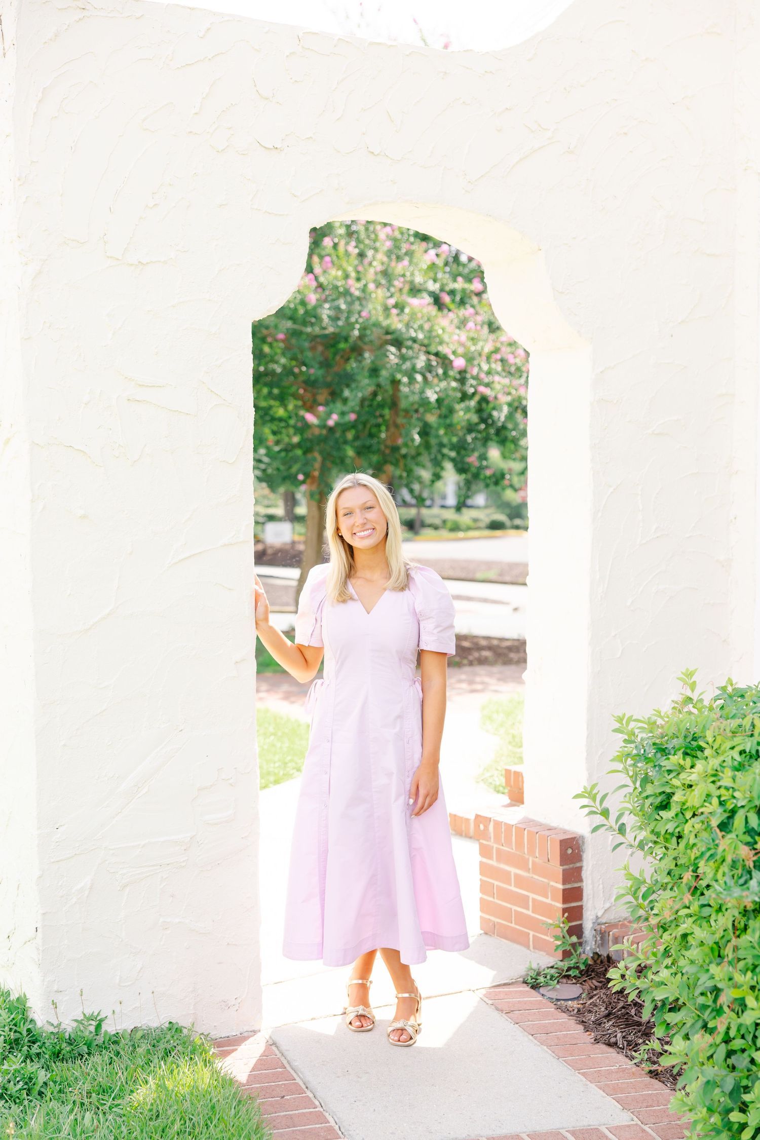A smiling blonde woman in lavender midi dress stands in a white arched doorway surrounded by greenery.