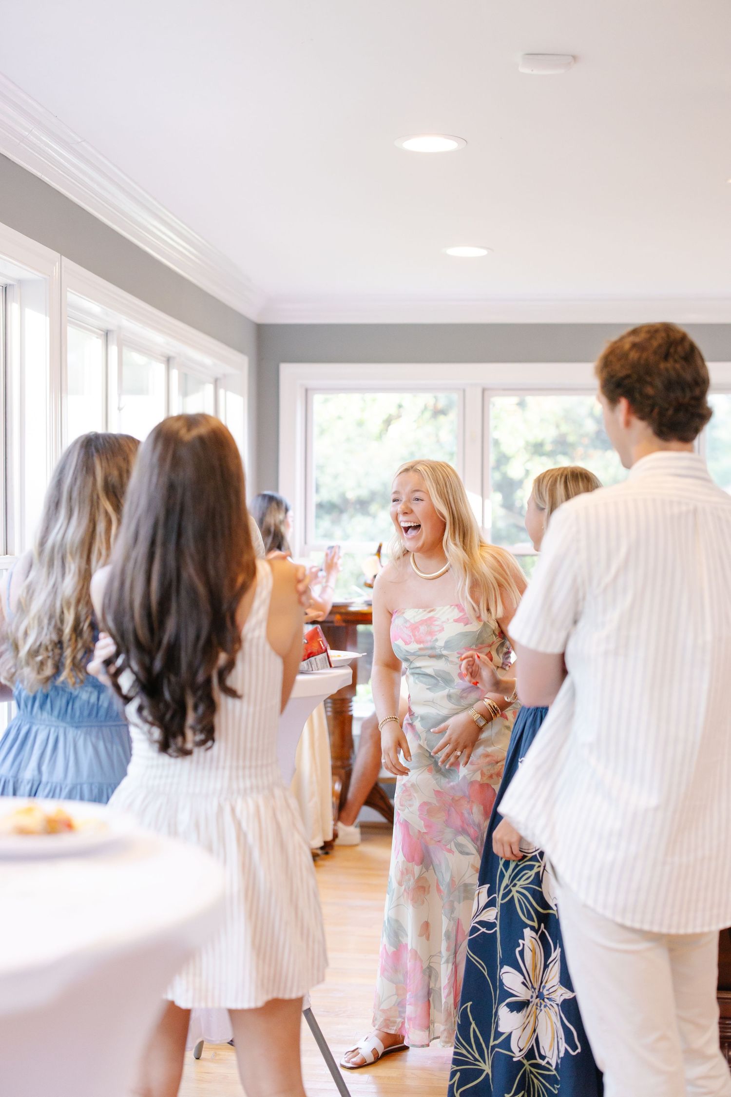 Laughing blonde woman in floral dress socializing with guests at a bright indoor celebration party.