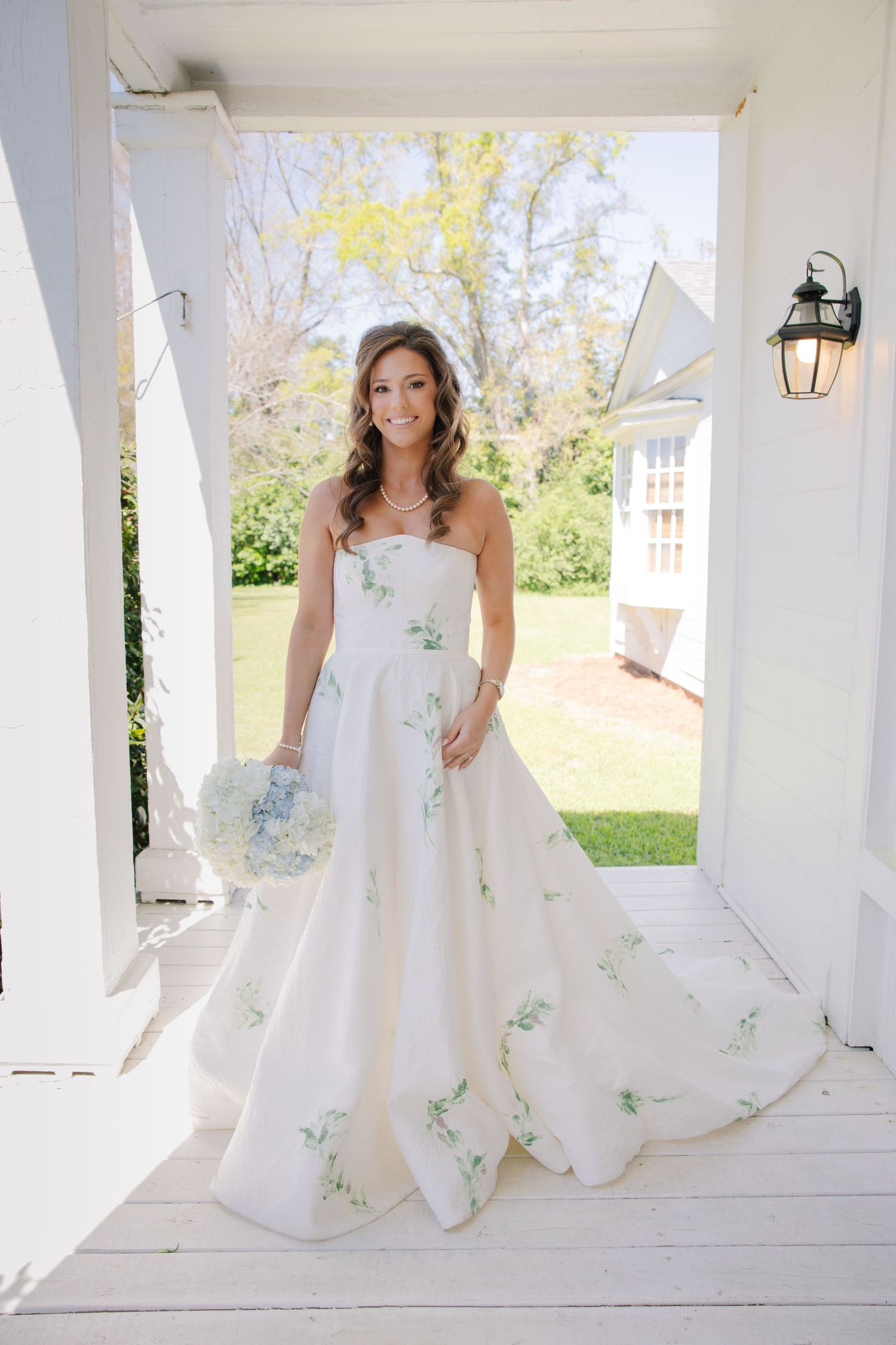 Bride in floral strapless ball gown holding hydrangea bouquet on white plantation porch.