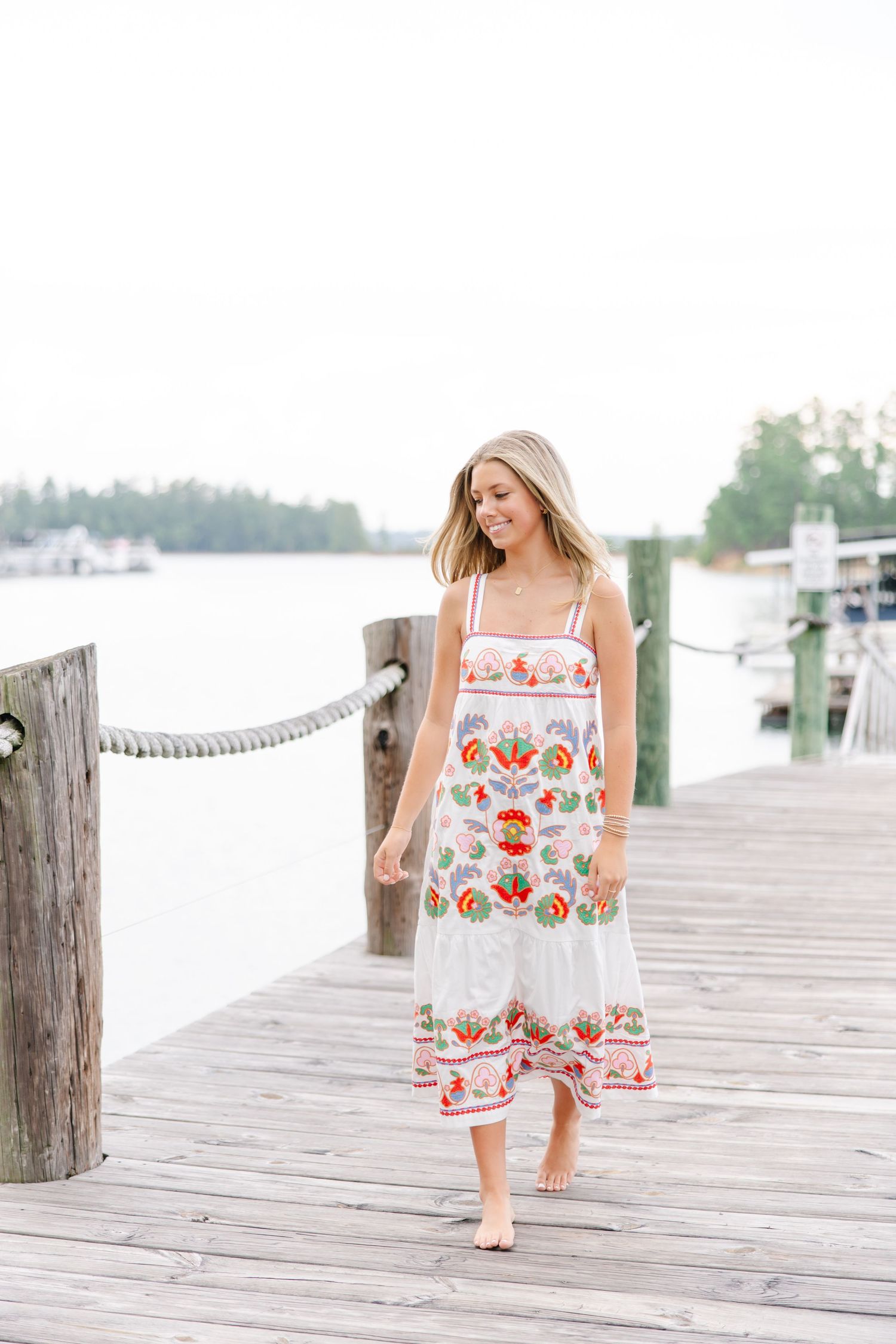 Blonde woman in white floral midi dress walking barefoot on wooden lakeside dock on overcast day.