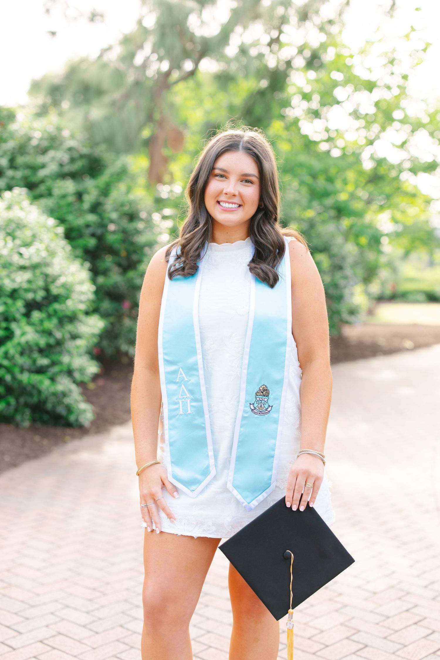 Happy graduate in white dress holding black cap with light blue stole on brick campus walkway.