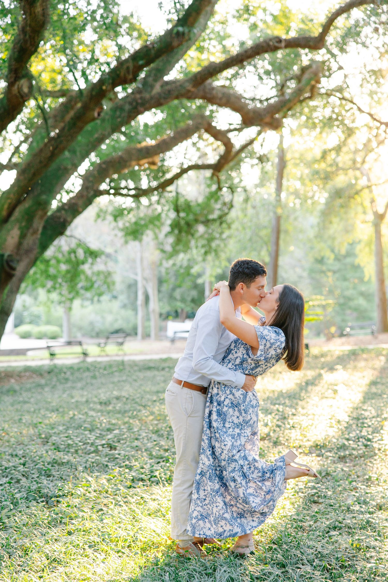 Couple shares romantic kiss under sprawling oak tree in golden hour sunlight at park.