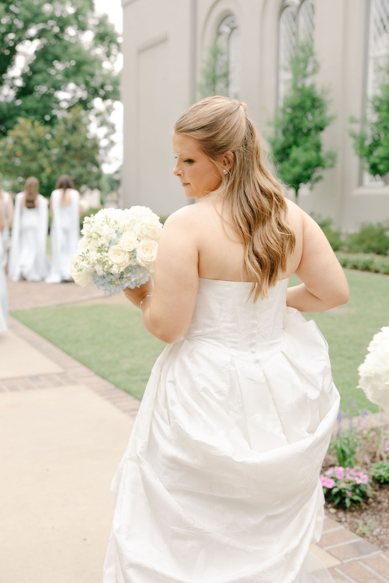 Bride in strapless white gown holds bouquet of white roses, walking with bridesmaids near a church.