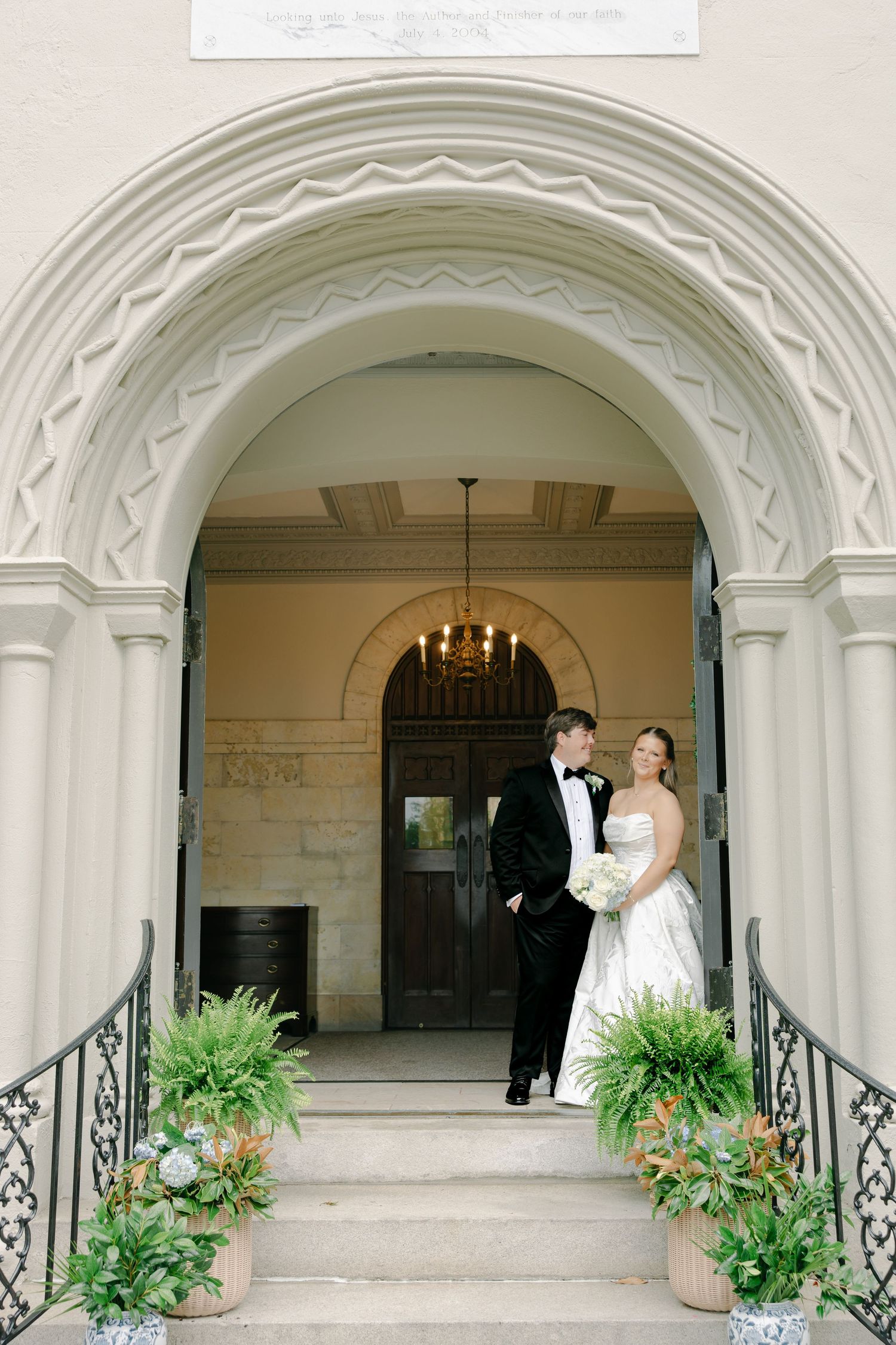 Bride and groom pose elegantly under ornate arch at church entrance with fern planters on stone steps.