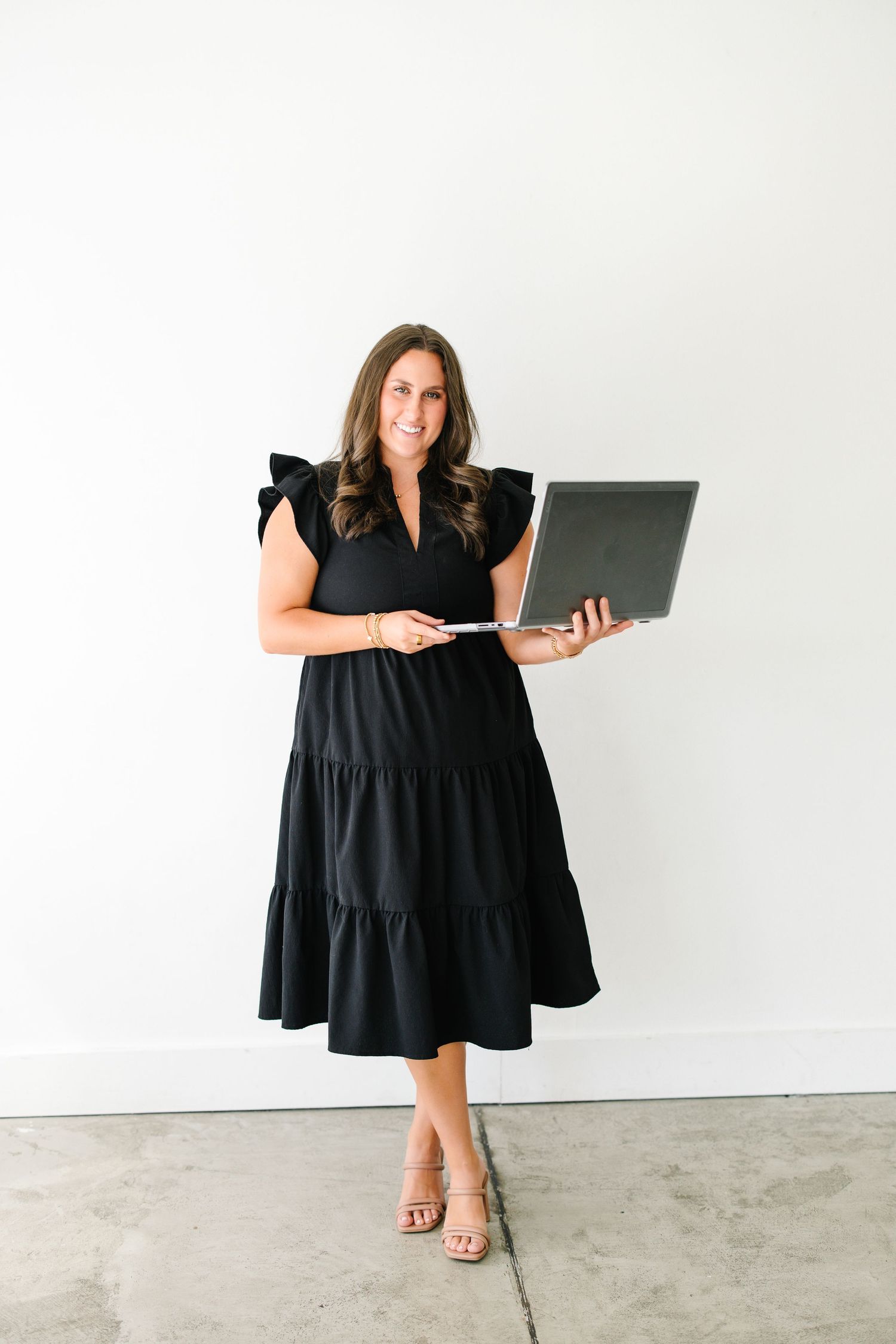 Woman in black ruffle dress holding laptop, smiling against white wall in professional business photo.