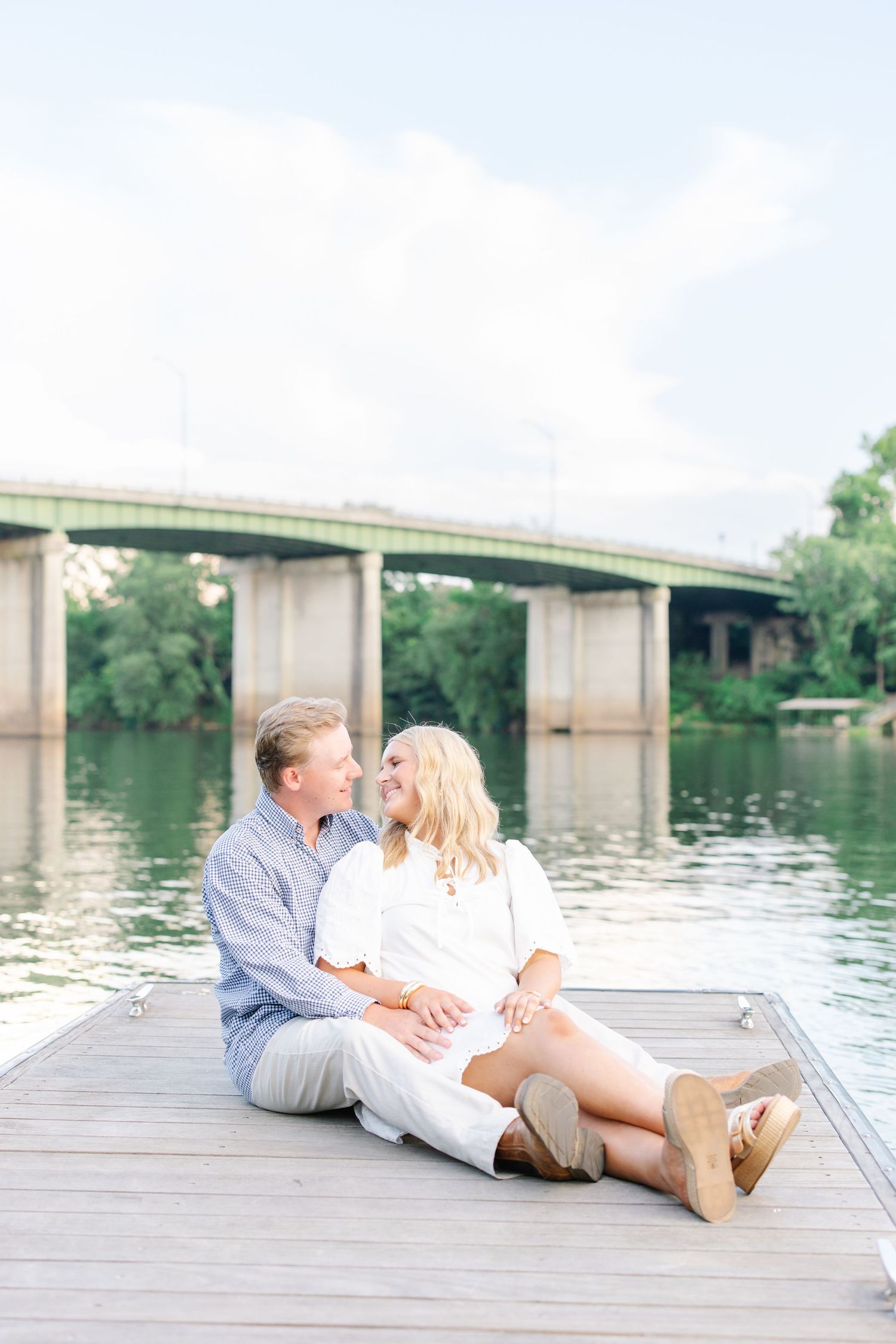 Couple sitting together on a wooden dock by a river smiling at each other with a bridge in the background.