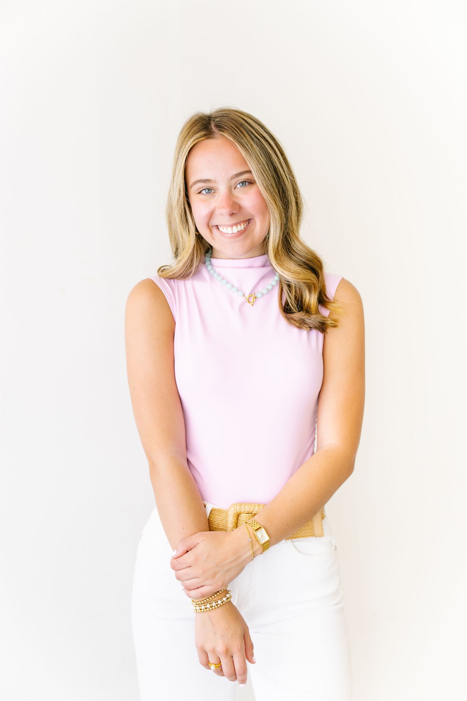 Smiling young woman in pink sleeveless top, white pants, and gold belt posing against a white background.