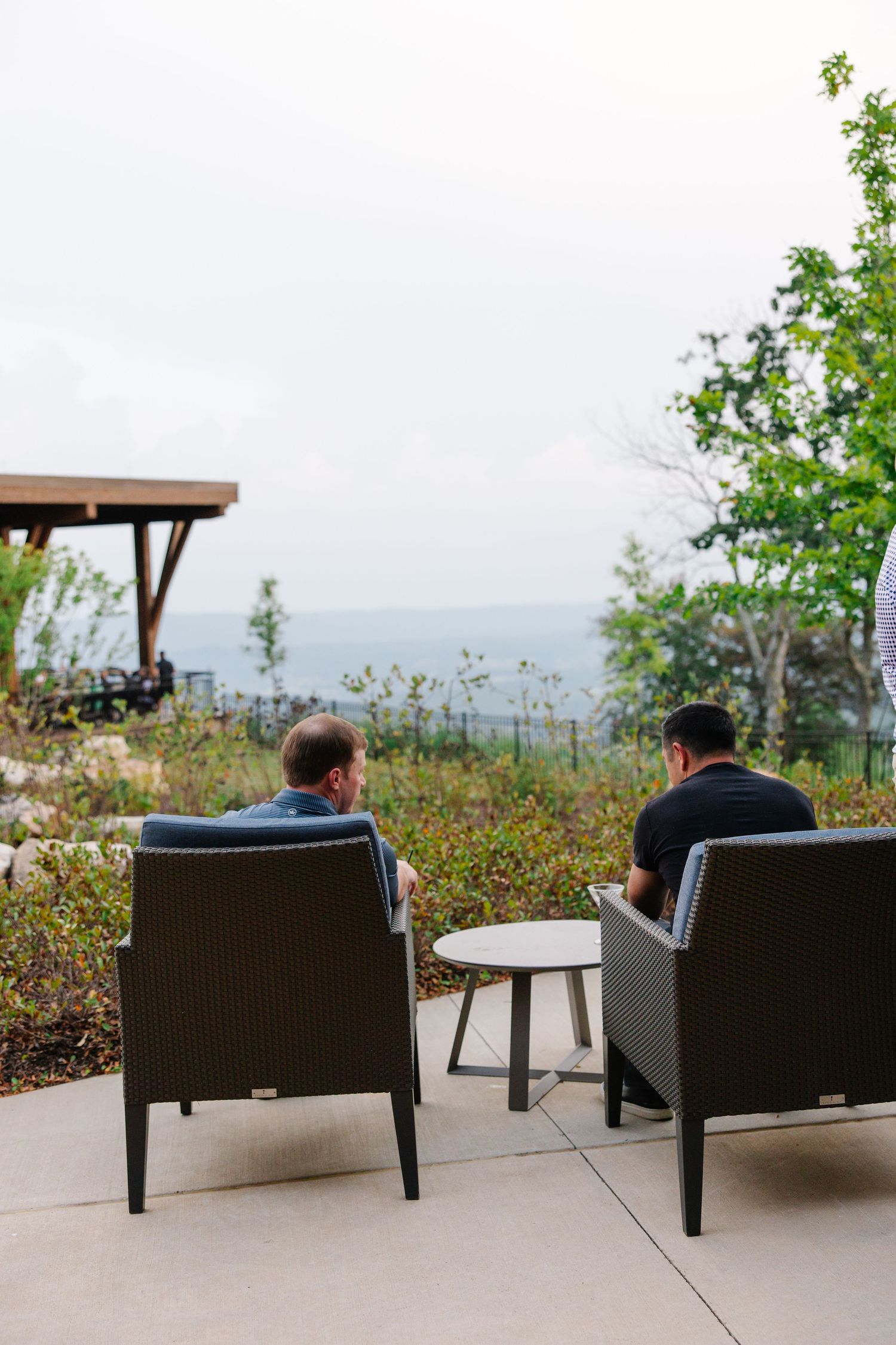 Two men relaxing in outdoor wicker chairs enjoying scenic mountain and valley views on a patio.