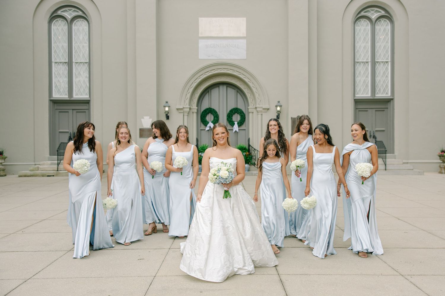 Bride in white ball gown walks confidently with bridesmaids in light blue satin gowns outside church.