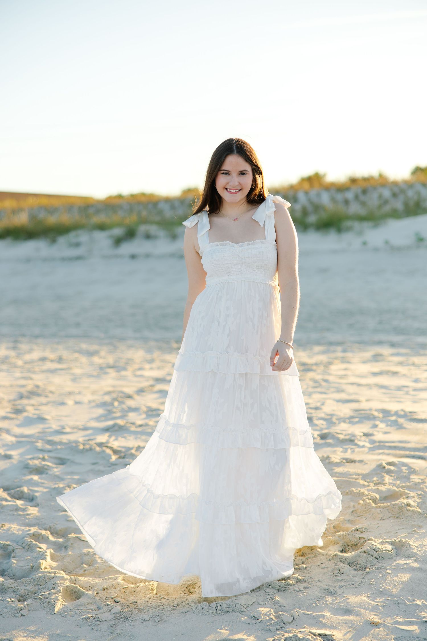 Smiling brunette woman in white tiered maxi dress posing on sandy beach at sunset.