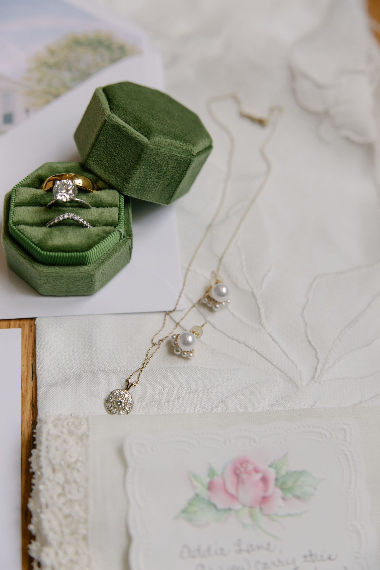 Green velvet ring box with diamond engagement ring, pearl necklace, and floral card on white linen.