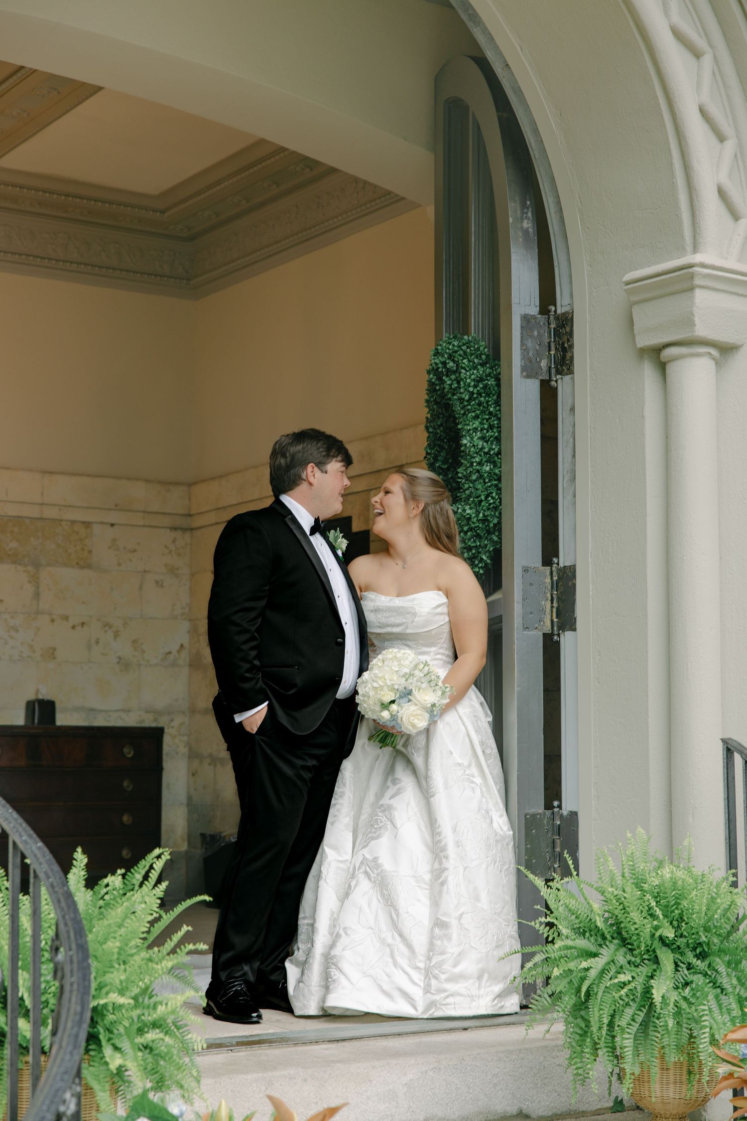 Bride and groom share a romantic moment on elegant venue steps, bride holding white bouquet in strapless gown, groom in black tuxedo.