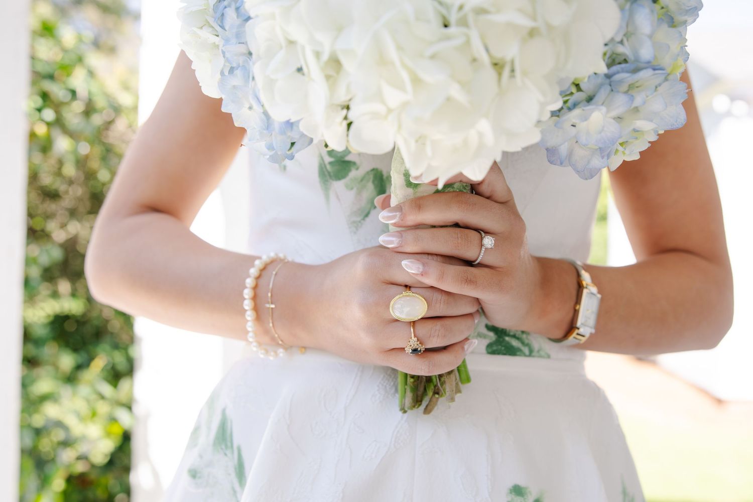Close-up of bride holding blue hydrangea bouquet wearing elegant rings and gold bracelet.