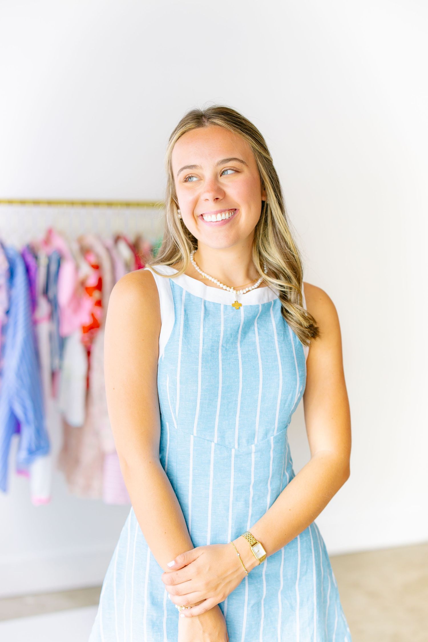 Smiling woman in blue striped sleeveless dress standing in front of a clothing rack with colorful garments.