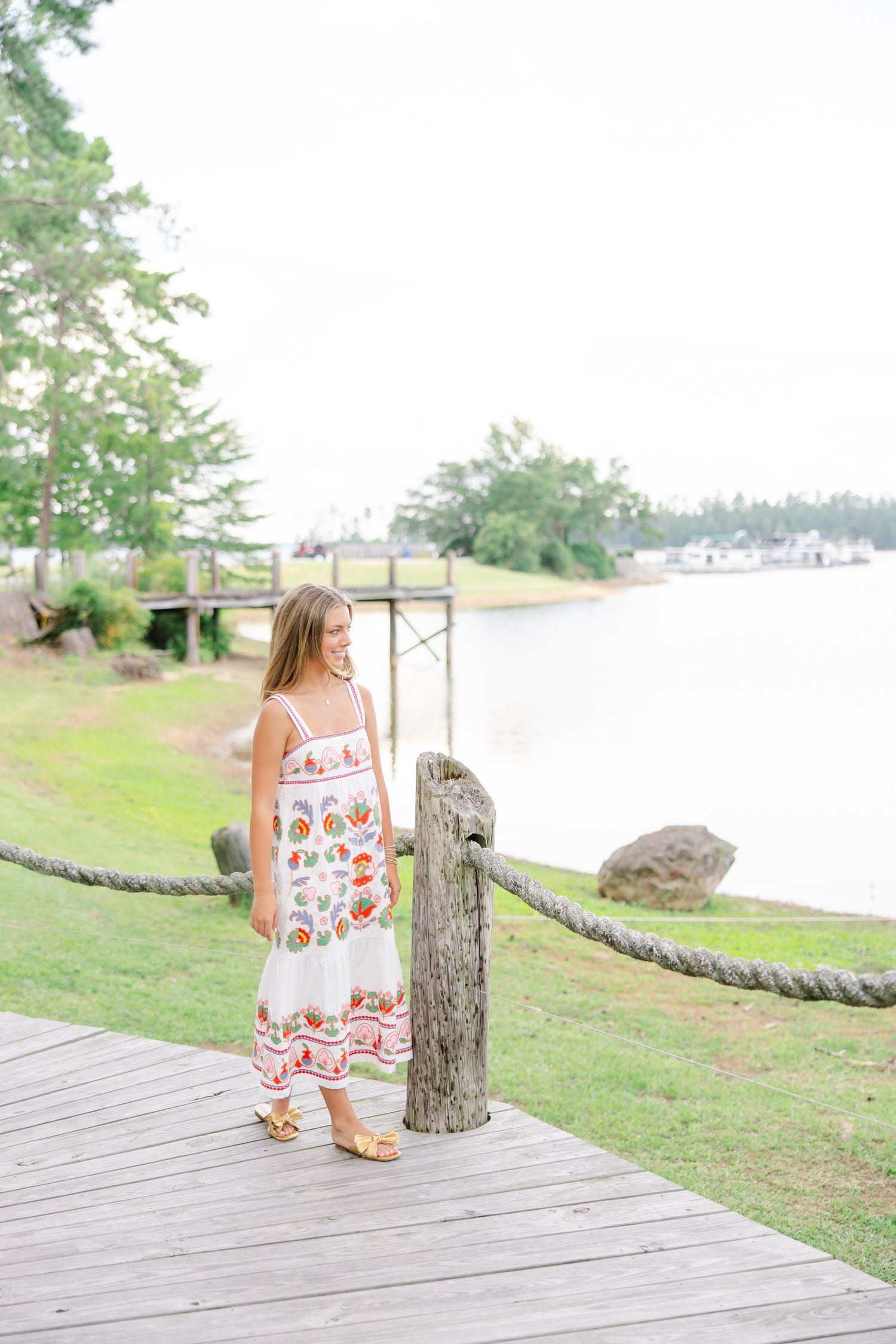 Young girl in white floral embroidered sundress standing on wooden dock by lakeside with rope railing.