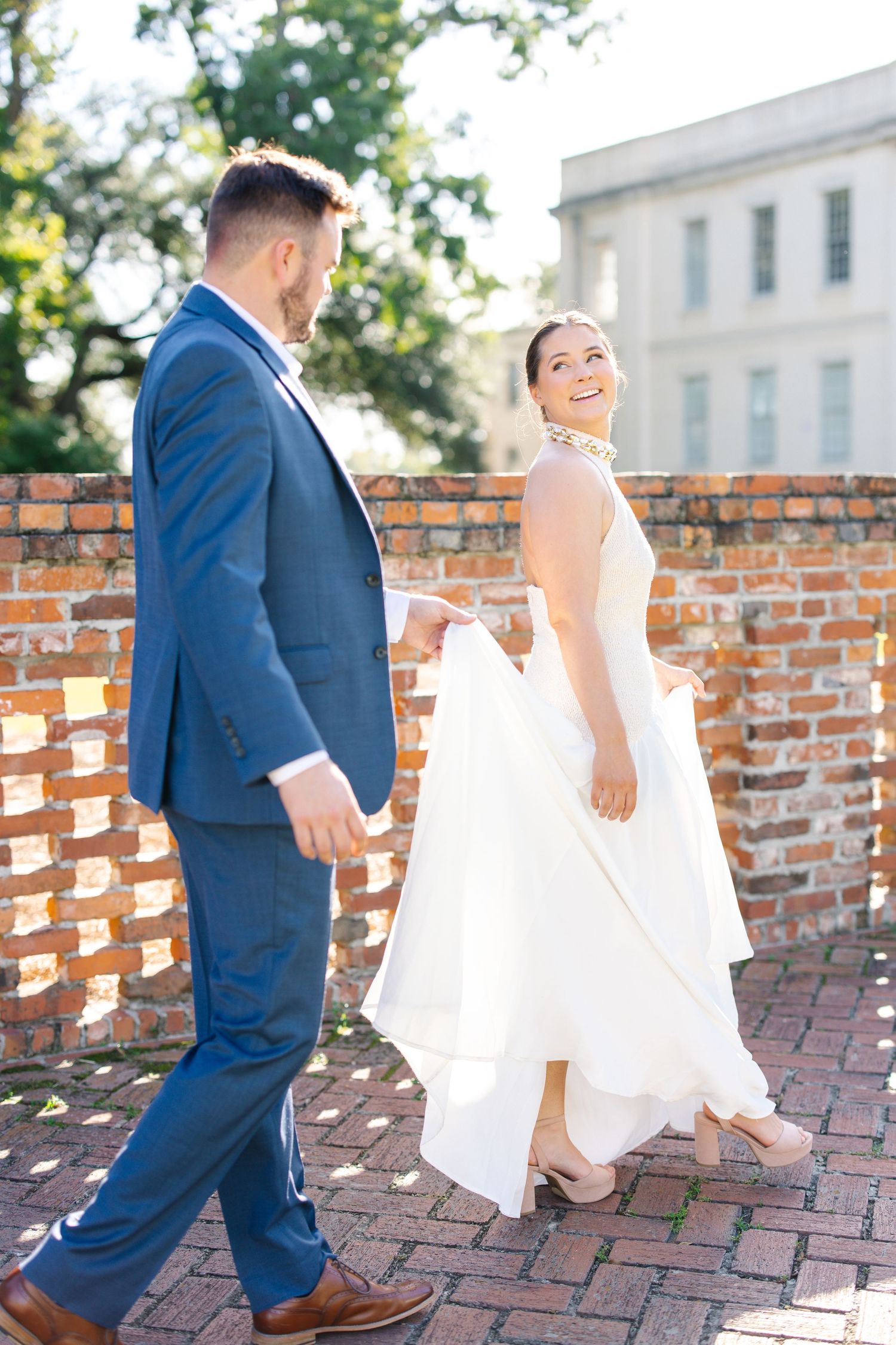 A smiling bride in white gown holds her dress while walking with groom in blue suit on brick path.