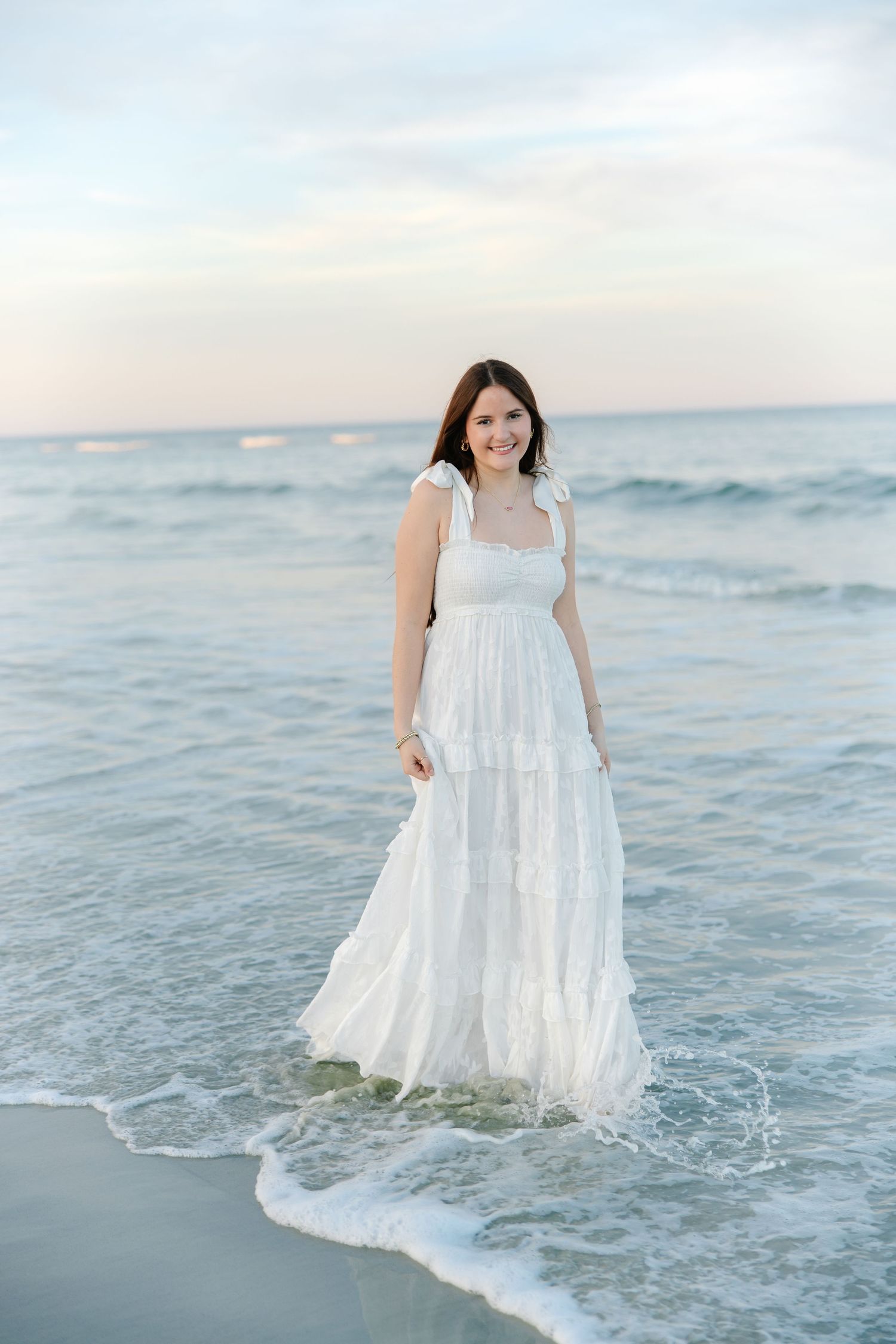 Smiling young woman in white tiered maxi dress stands in ocean waves at sunset beach.