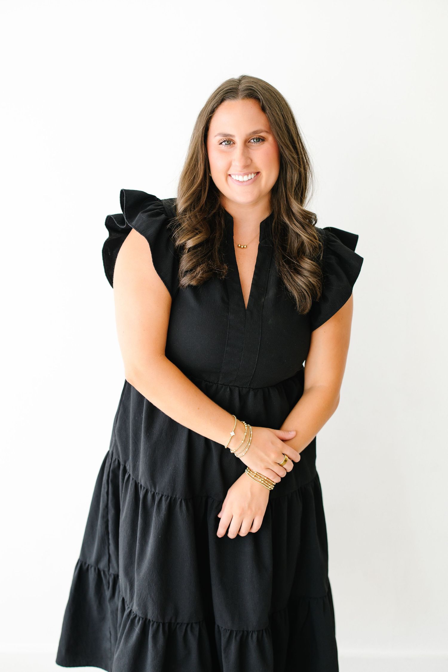 Smiling brunette woman in black ruffled dress with gold bracelets poses against white background.