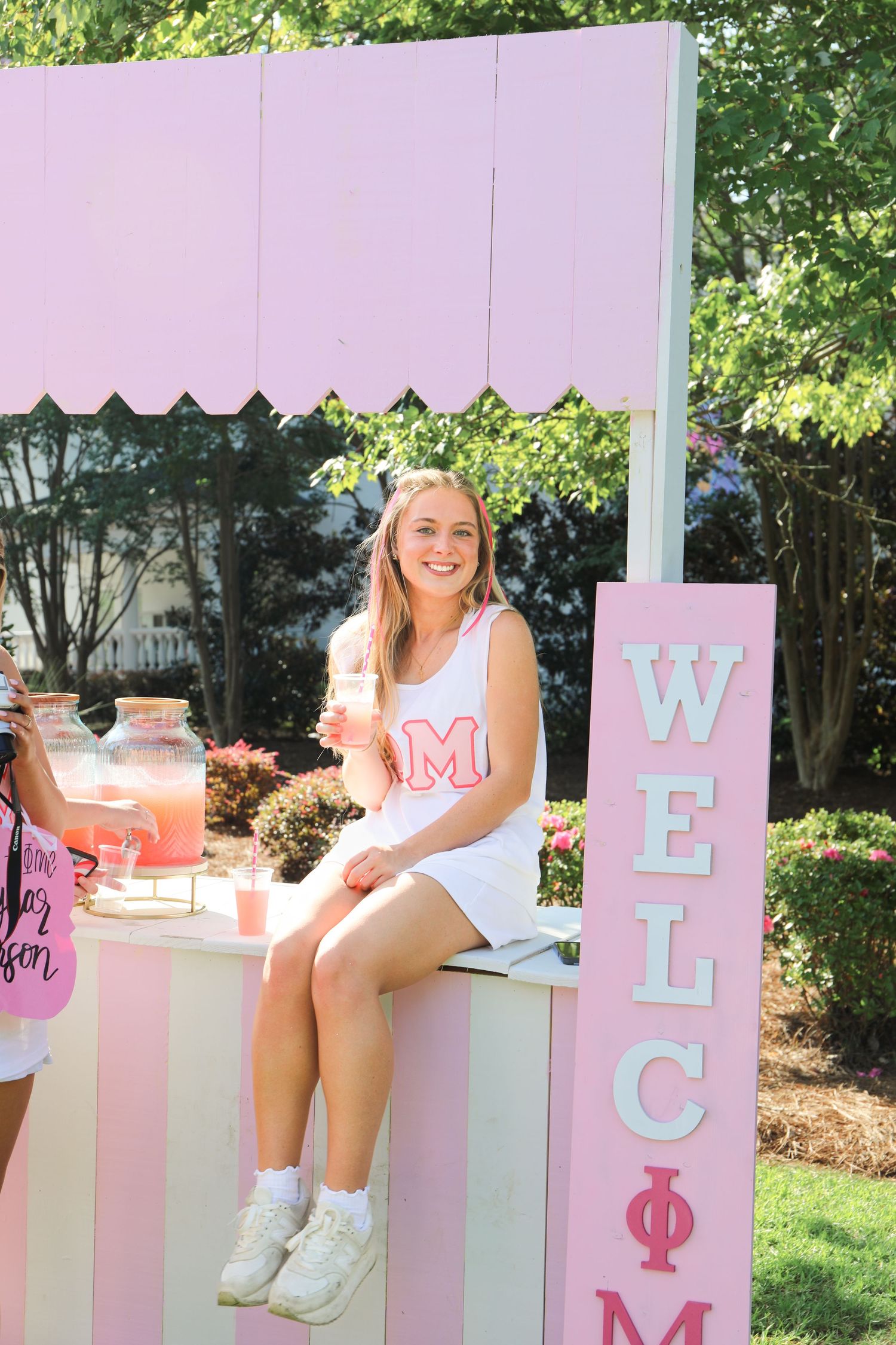 Smiling blonde woman sitting on pink lemonade stand booth at a Phi Mu sorority welcome event outdoors.