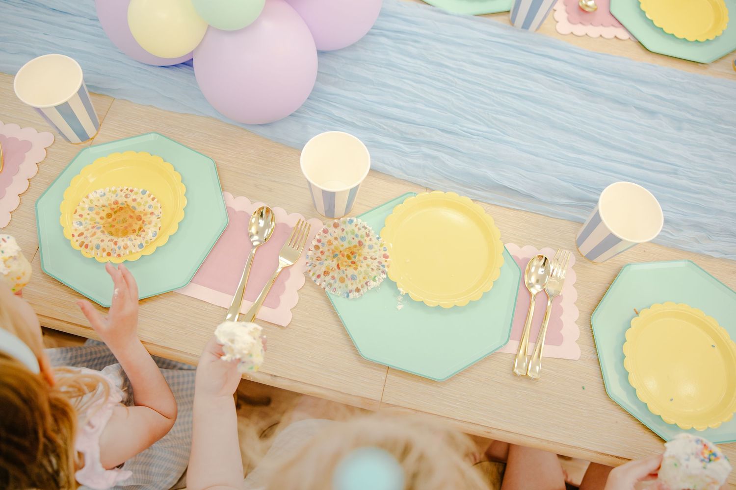 Overhead view of a pastel birthday party table with mint and yellow plates, gold cutlery, and striped cups.