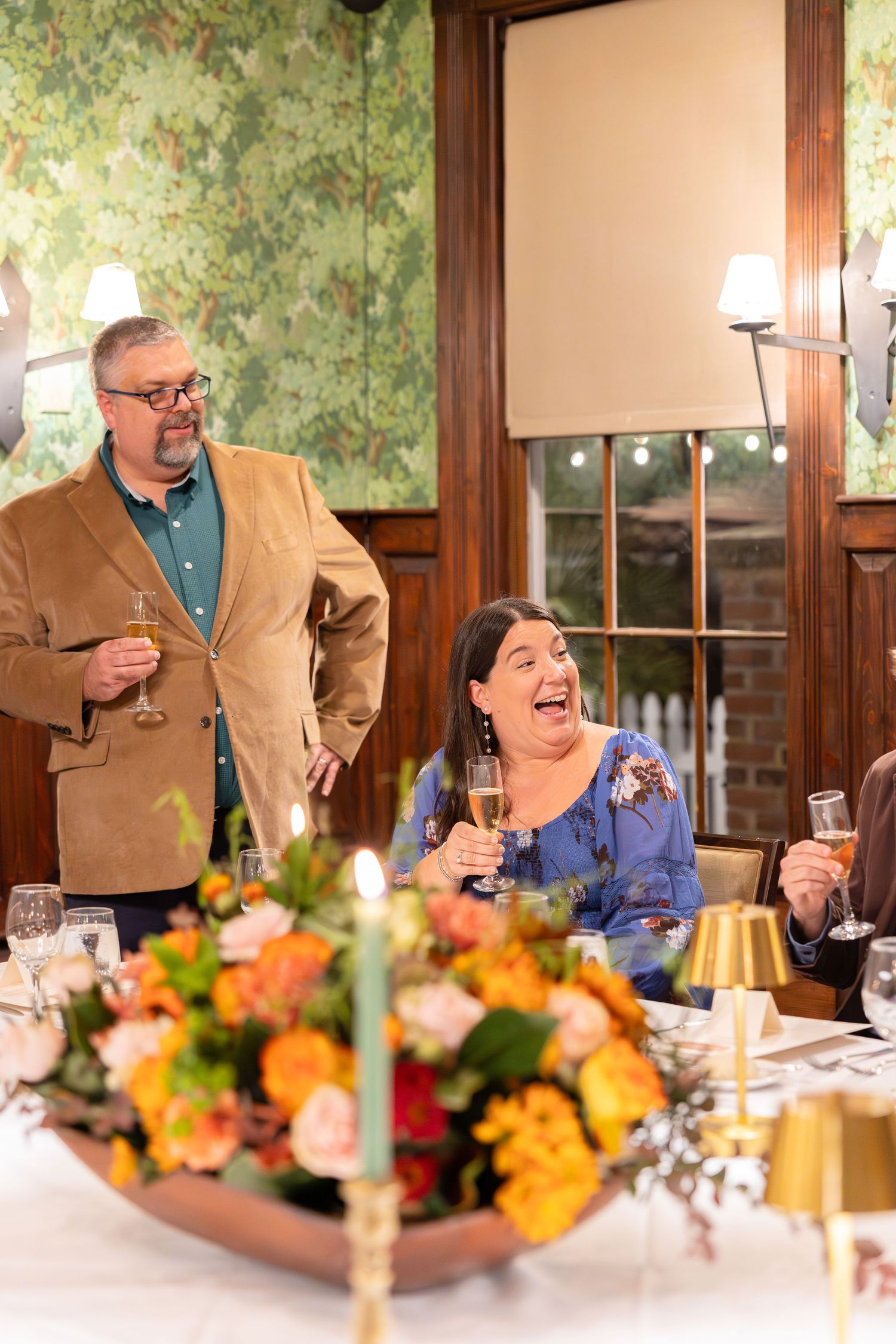Man and woman laughing and raising champagne glasses during a celebration dinner with orange floral centerpiece.