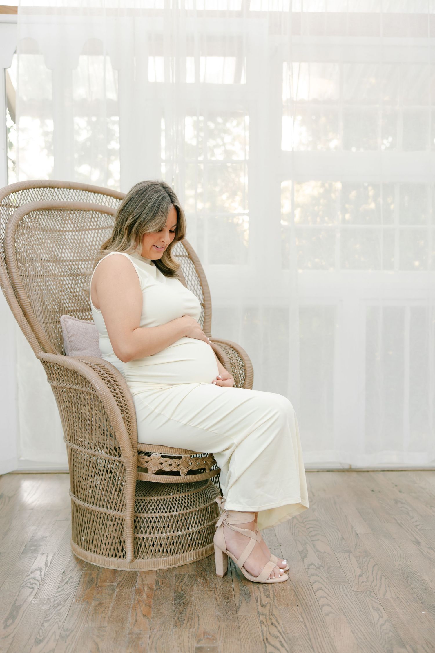 Pregnant woman in white outfit sitting in wicker chair holding baby bump.