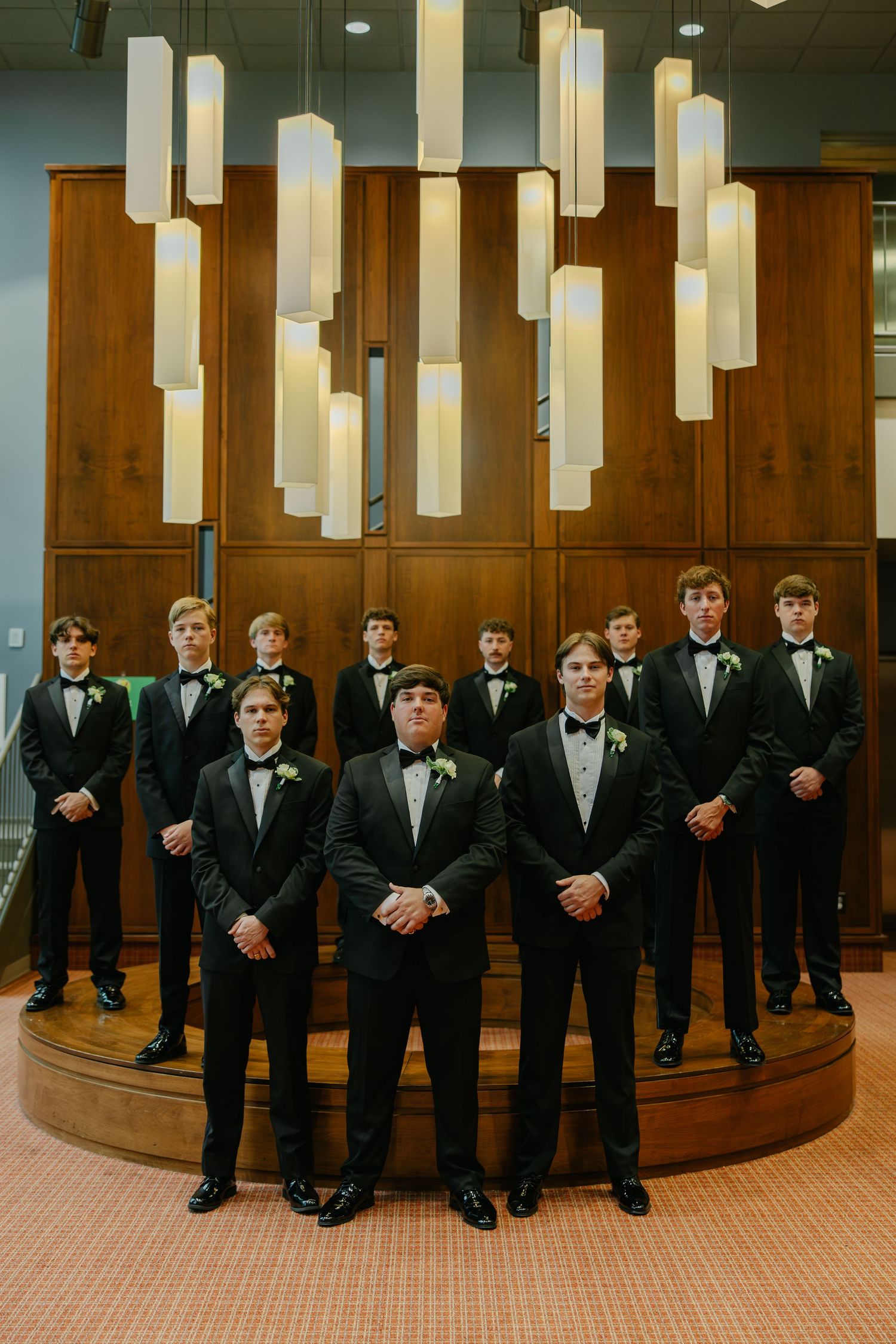 Groom and groomsmen in black tuxedos pose formally under modern pendant lights in an elegant indoor venue.