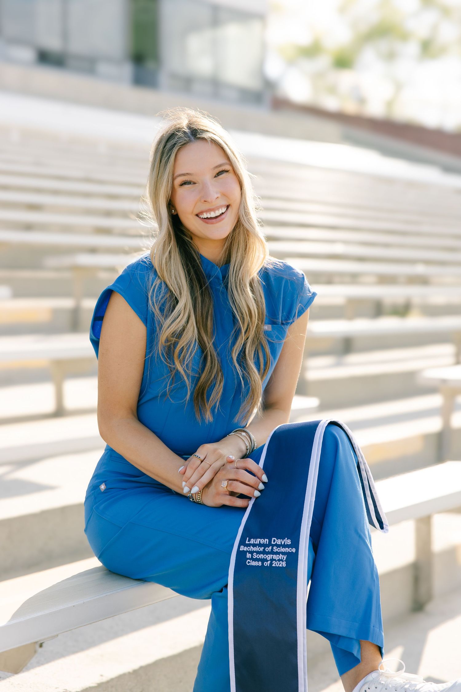 Smiling blonde graduate sits on bleachers in blue outfit holding her personalized graduation stole outdoors.