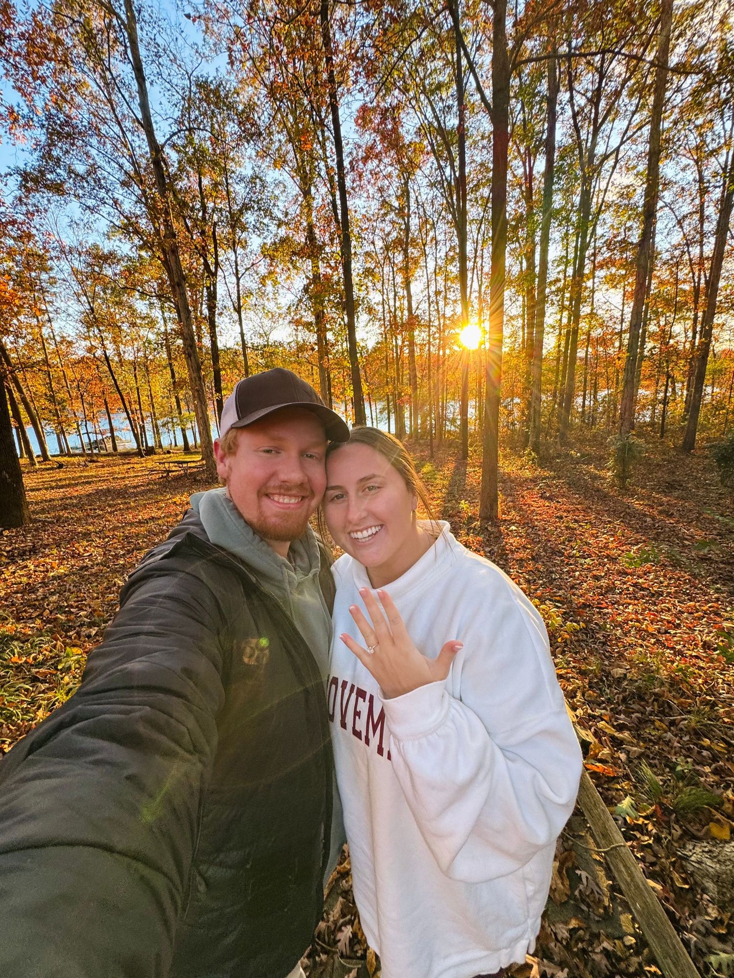 Couple taking a selfie in autumn woods at sunset showing off an engagement ring among colorful fall foliage.