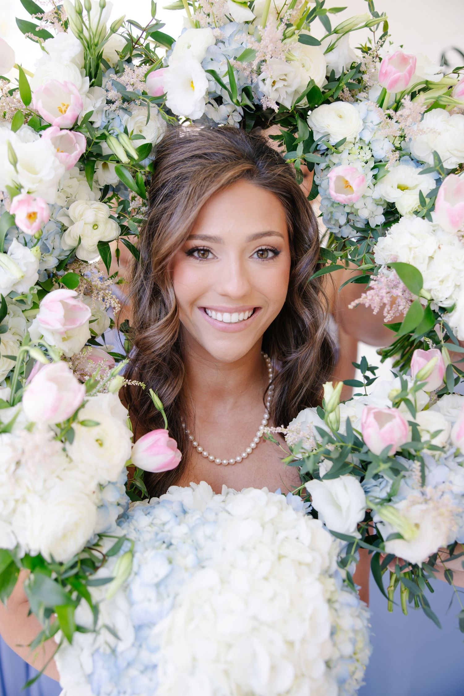 Smiling bride surrounded by lush white and pink floral arrangements wearing pearl necklace.