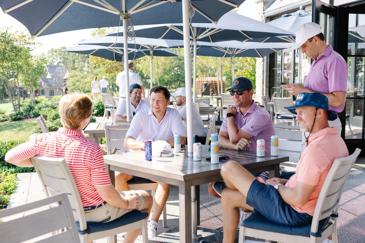 Group of golfers in polo shirts socializing at an outdoor patio table under umbrellas.