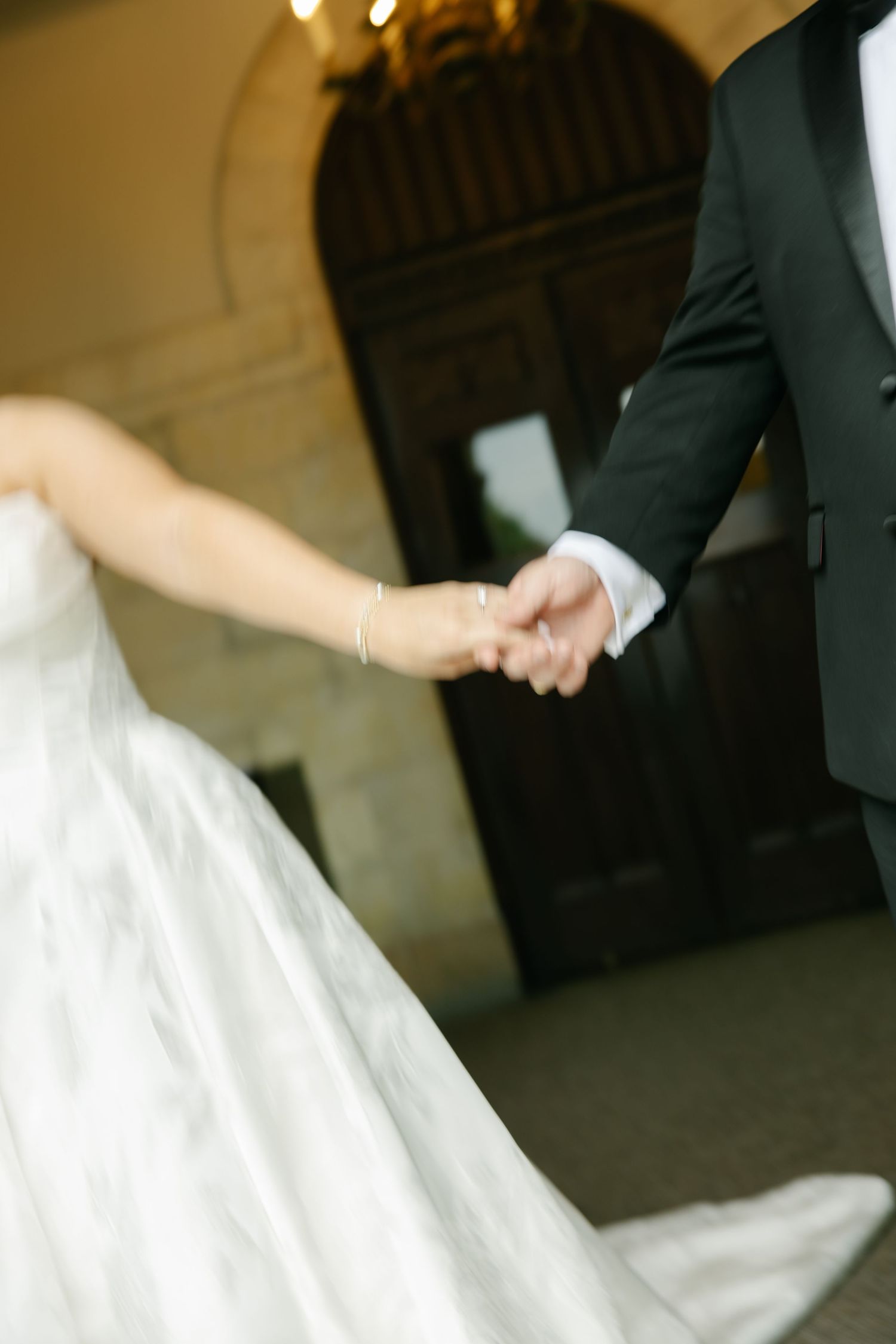 Bride and groom holding hands in elegant wedding attire, white gown and black tuxedo, near ornate wooden doors.